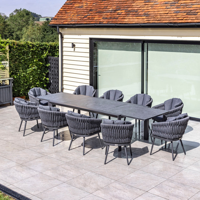 Outdoor patio featuring the Palma 10 Seat Rope Extending Dining Set in Grey with cushioned chairs on a tiled surface, beside a house with glass doors. The dining set boasts durable aluminium frames for longevity and style.