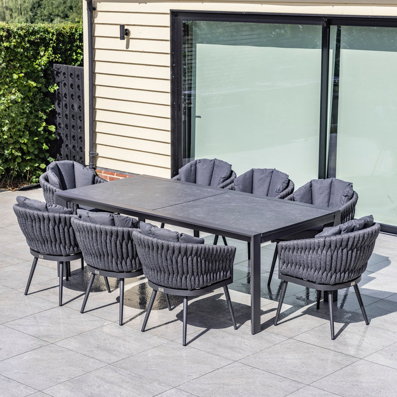 Outdoor patio set with a rectangular table and eight cushioned chairs on a tiled floor, featuring the Palma 8 Seat Rope Extending Dining Set in Grey by Harbour Lifestyle. Built next to a house with large windows, this high-quality aluminum ensemble promises durability and style.