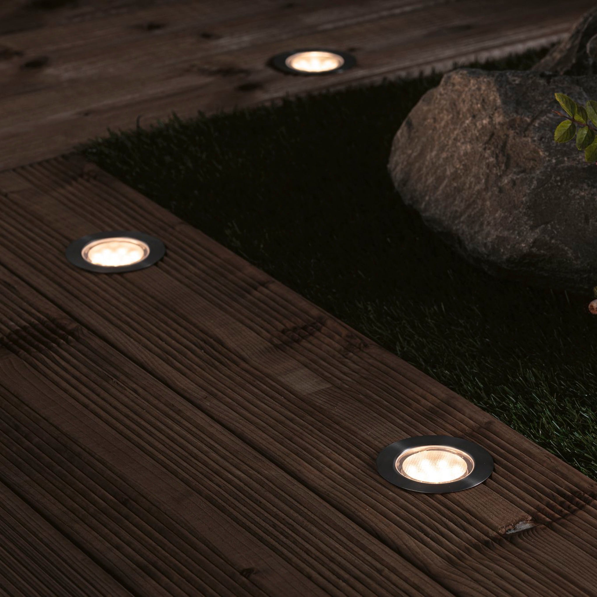 With the Paulmann - Plug & Shine Outdoor 2W LED Recessed Floor Individual Eco Spotlight in Silver, a wooden terrace is beautifully illuminated at night, casting a warm glow across the outdoor space. The serene backdrop is elegantly enhanced by grass and rocks.