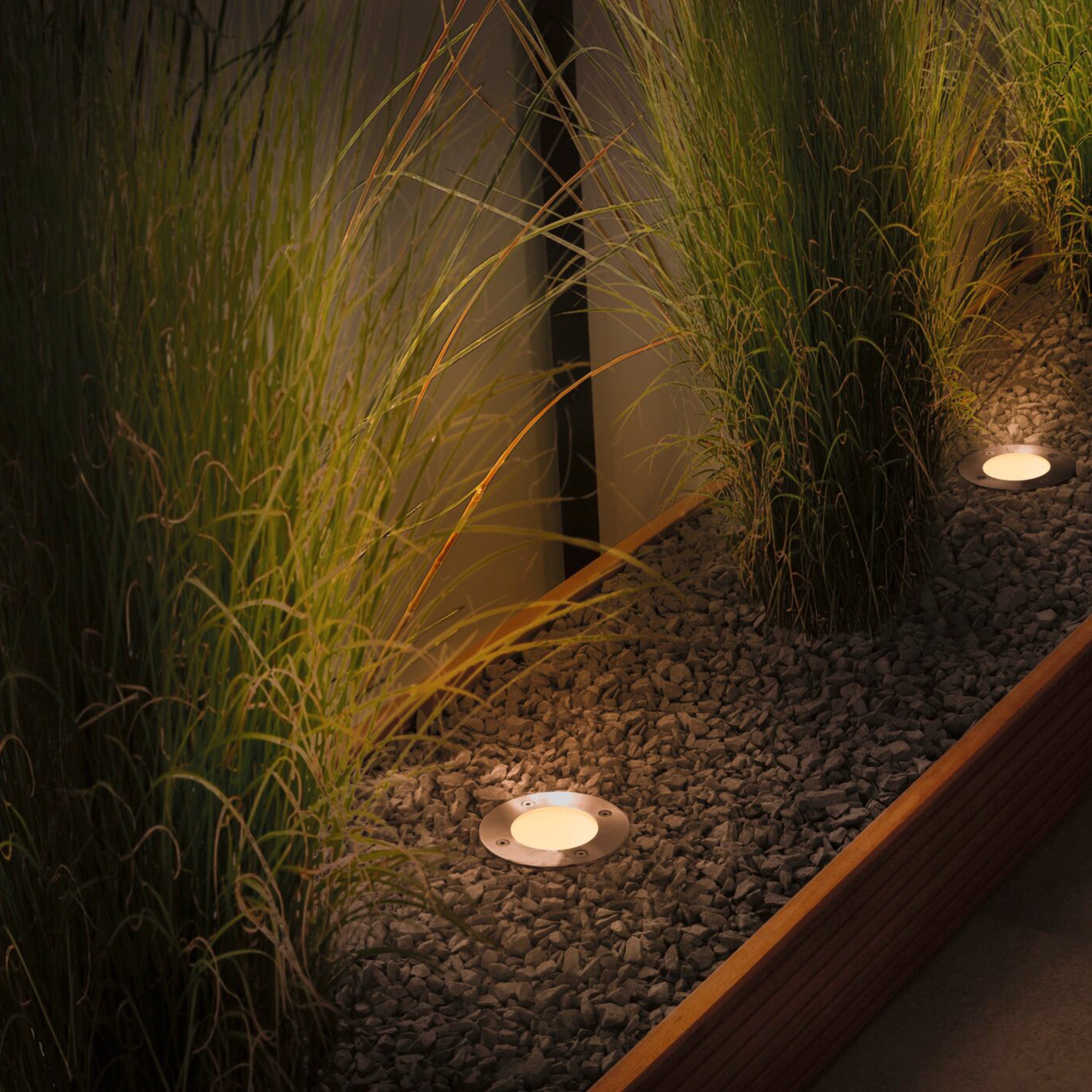 A garden pathway is beautifully illuminated with Paulmann's Plug & Shine Insect Friendly Outdoor 4.5W LED Recessed Floor Individual Spotlights in Anthracite, casting warm white light over the tall grasses on gravel while keeping insects undisturbed.