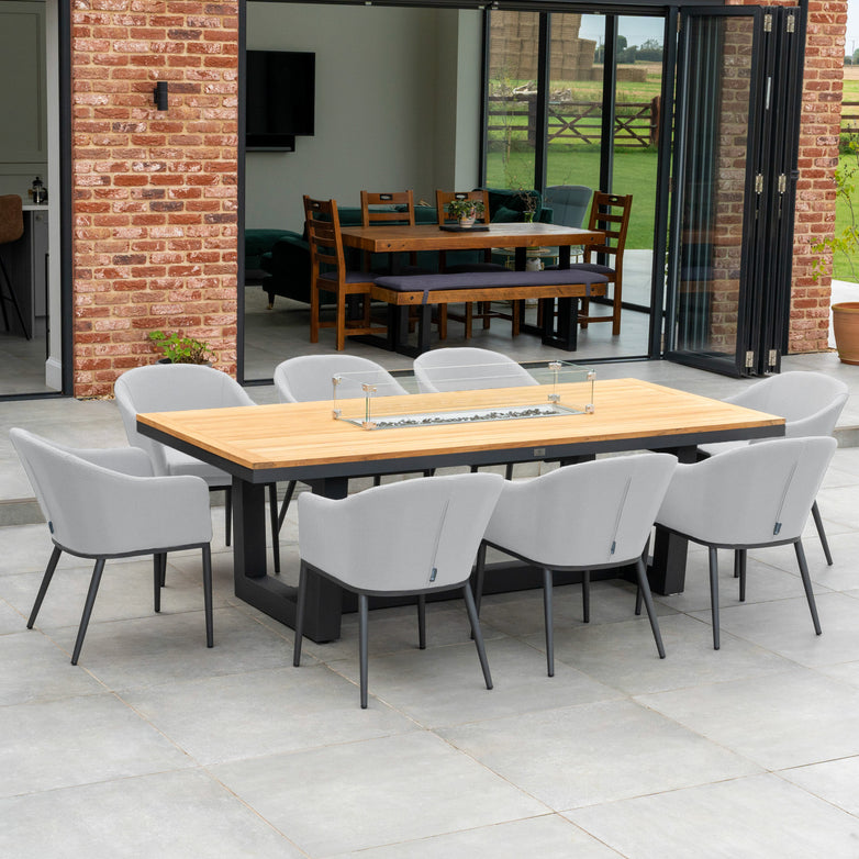 Outdoor dining area featuring the Luna 8 Seat Outdoor Fabric Rectangular Teak Firepit Dining Set in Oyster Grey, placed on a patio with brick walls and folding doors for added comfort and ambiance.