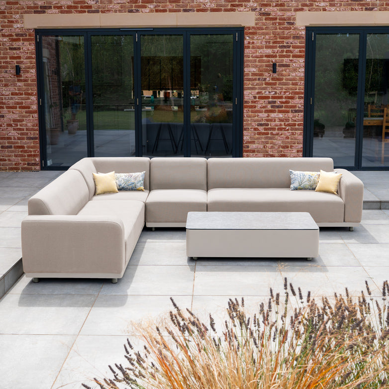 The Luna Outdoor Fabric Large Corner Group Set with Coffee Table in Fawn, featuring an aluminium frame, is arranged on a patio before glass doors and a red brick house.