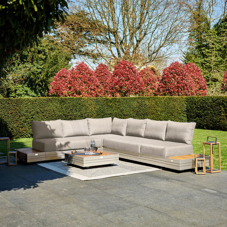Outdoor patio with the Panama Luxury Outdoor L-Shape Corner Group Set in Latte featuring a gray sectional sofa and low coffee table on a paved surface, surrounded by greenery and red bushes.