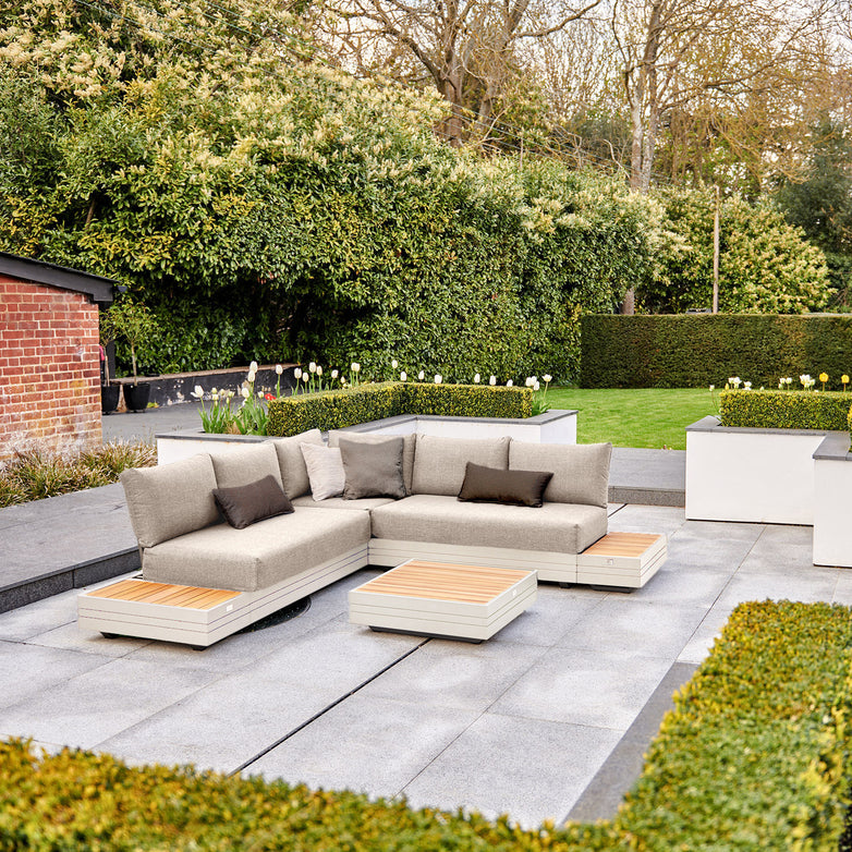 Outdoor patio with the Panama Luxury Outdoor Corner Group Set in Latte featuring a gray sectional sofa, wooden-top tables crafted from sustainable teak, and neatly trimmed garden bushes and grass in the background.