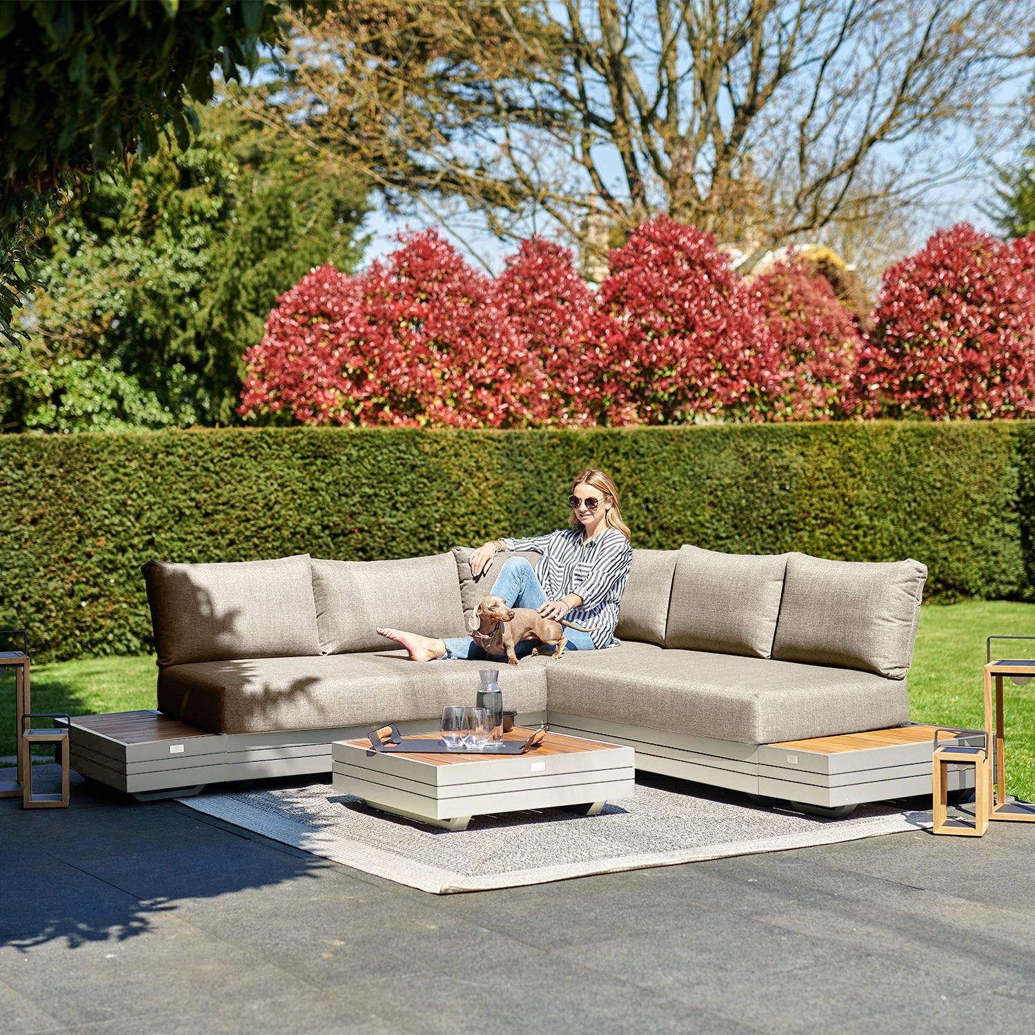 A woman sits on an outdoor sectional sofa with a dog, surrounded by greenery and red shrubs, on a stone patio. The Panama Luxury Outdoor Corner Group Set in Latte by Harbour Lifestyle adds a touch of elegance to the serene scene.