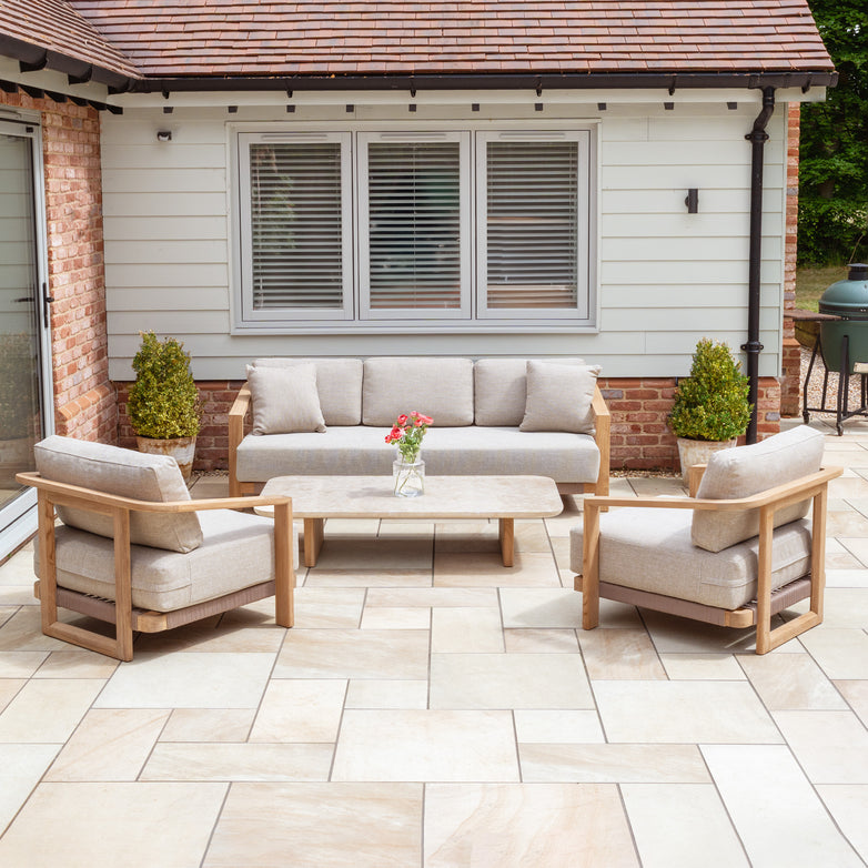 Patio with a Tivoli 3 Seat Sofa Set in Fawn and marble coffee table, accented by beige cushions and teak wood, surrounded by potted plants with a house in the background.