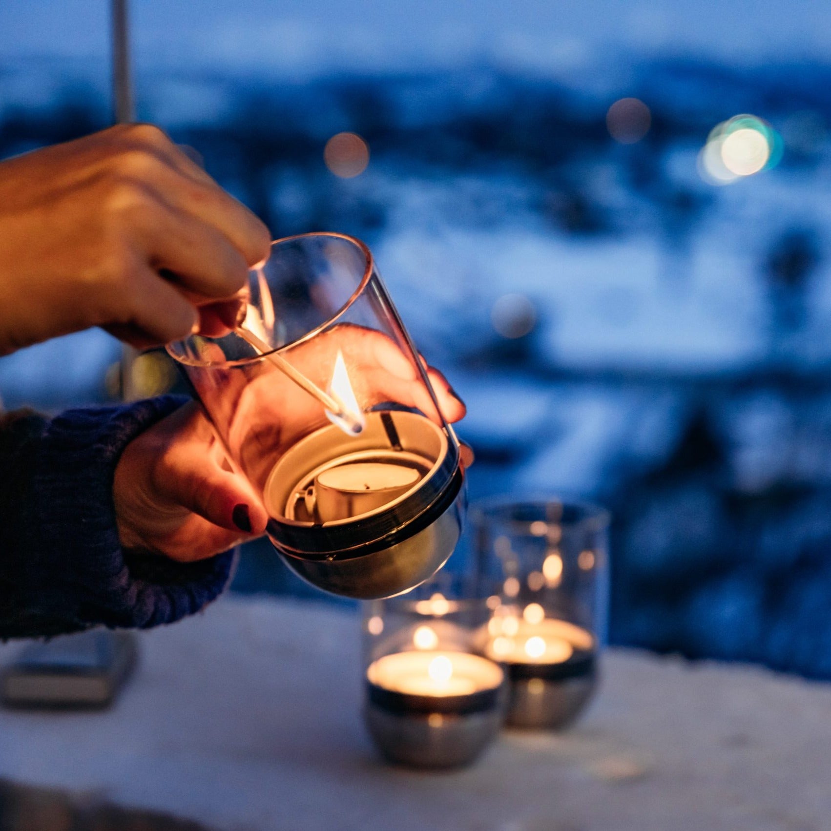 Hands lighting a candle with a matchstick; snowy landscape and more lit candles in the blurry background, possibly those of the elegant Höfats GRAVITY CANDLE Lantern, perfect for both indoor and outdoor use.