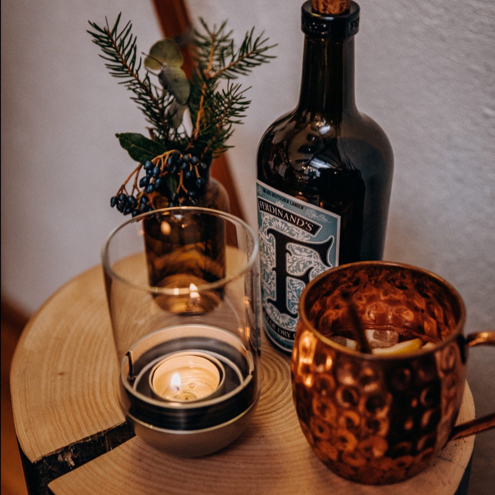 A lit Höfats GRAVITY CANDLE Lantern, a dark bottle, a metal mug with a slice of lemon, and a small plant rest on a wooden surface.