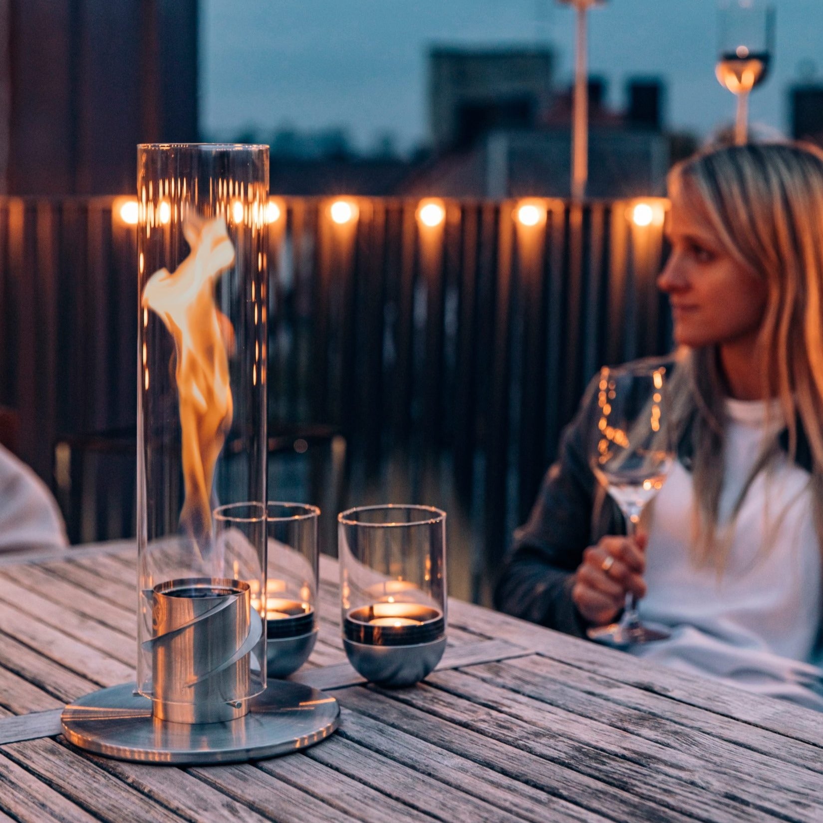 A woman holding a wine glass sits at an outdoor table with a Höfats GRAVITY CANDLE Lantern and candles in the evening.