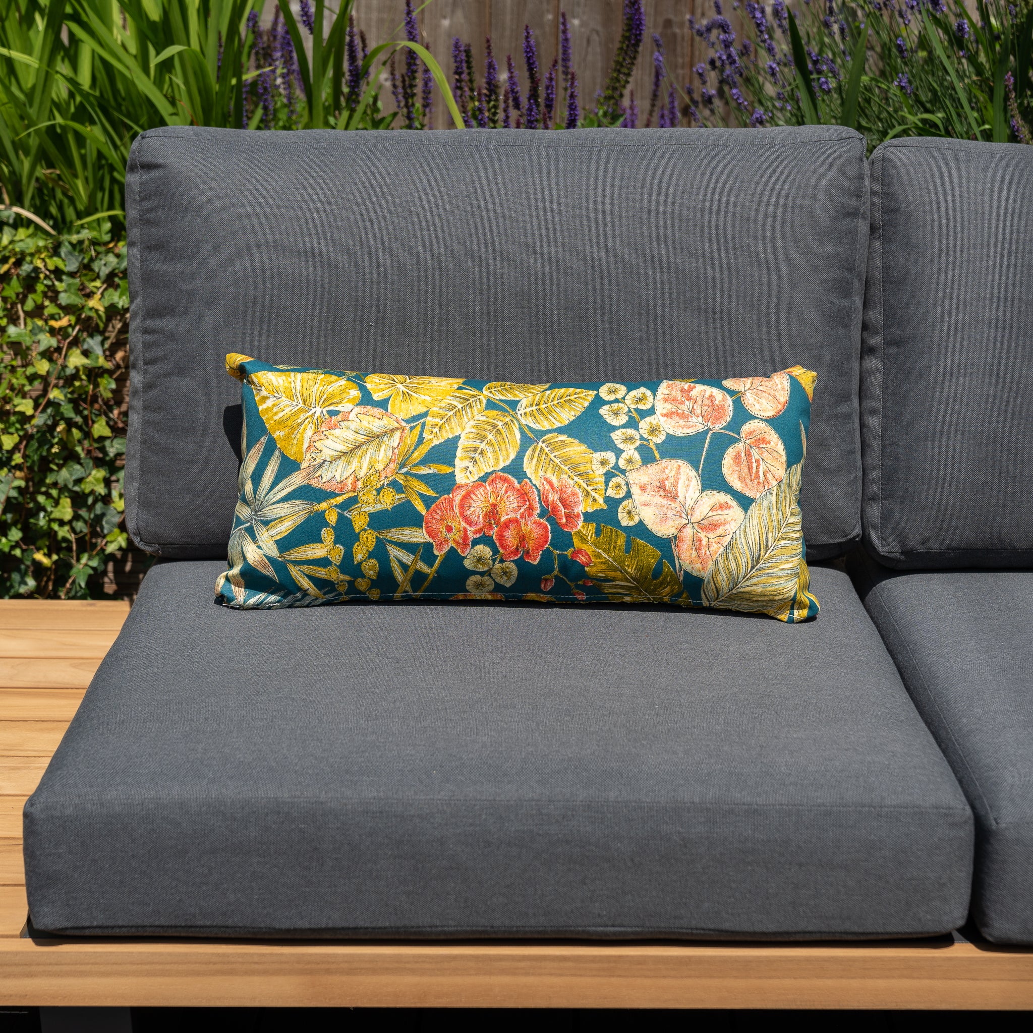 An Acrisol Bouquet Petrol Bolster Scatter Cushion (60cm x 30cm) in vibrant florals adorns a gray outdoor sofa, complemented by lush green plants and purple flowers in the background, showcasing a range of exquisite solution-dyed acrylic fabrics.