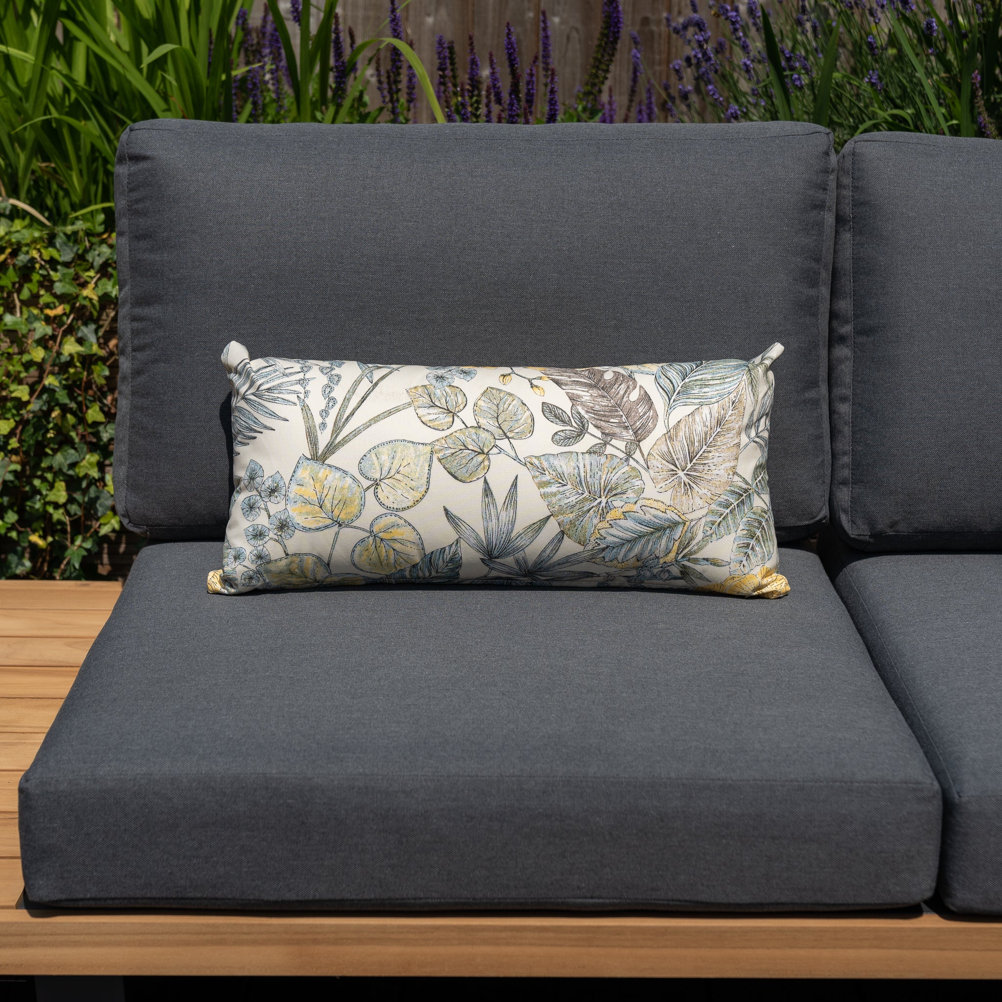 A gray outdoor sofa with a high-quality Acrisol Bouquet Neutrals Bolster Scatter Cushion, featuring floral and leaf patterns on solution-dyed acrylic fabric, situated on a wooden deck in a custom outdoor space.