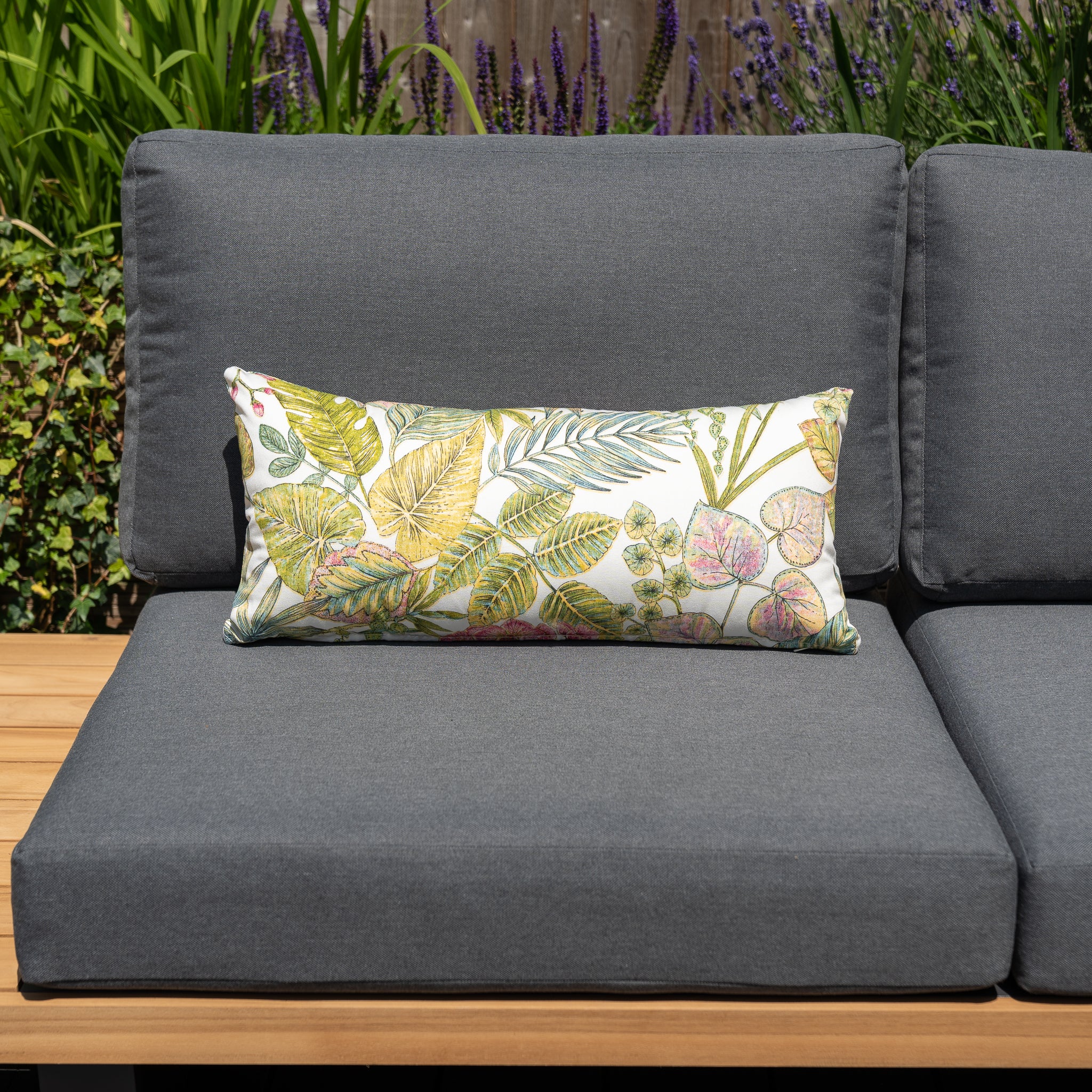 A gray outdoor sofa adorned with a floral-patterned cushion on the seat, accentuated by the Acrisol Bouquet Multi Bolster Scatter Cushion (60cm x 30cm) and matching outdoor scatter cushions, creates a vibrant and charming garden setup amidst greenery and purple flowers.