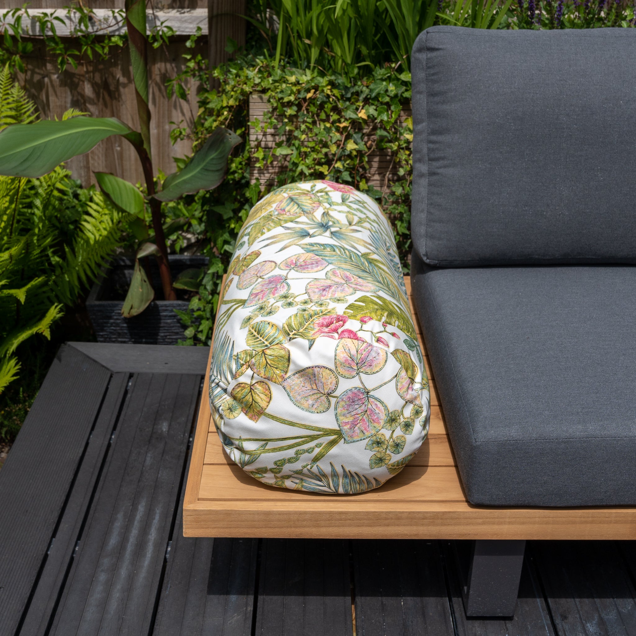 Outdoor seating area with solution-dyed acrylic cushion storage bag, gray cushion, and the vibrant Acrisol Bouquet Multi Large Round Bolster Floor Cushion - 72cm x 35cm on a wooden deck amidst lush plants.