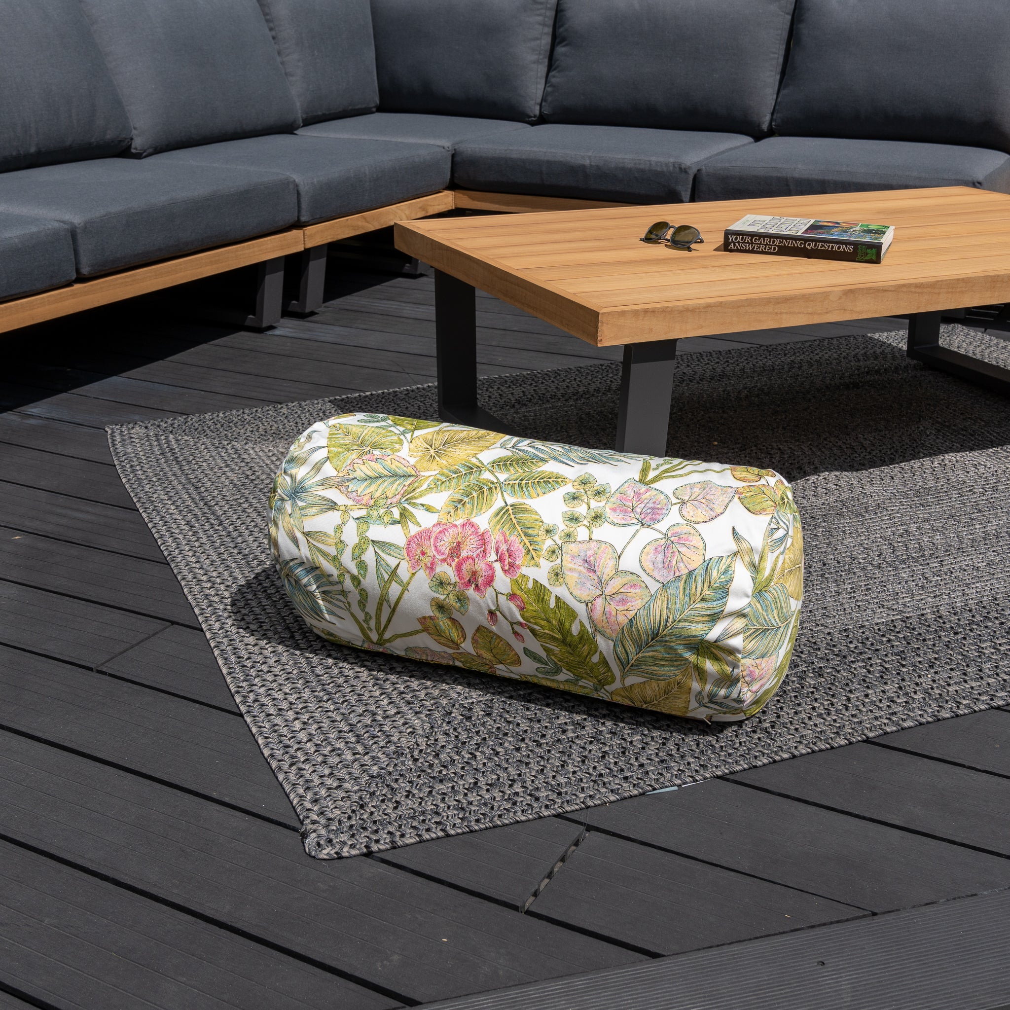 An Acrisol Bouquet Multi Large Round Bolster Floor Cushion (72cm x 35cm) on a gray outdoor rug beside a wooden coffee table and gray sectional sofa, accented with bright outdoor scatter cushions.
