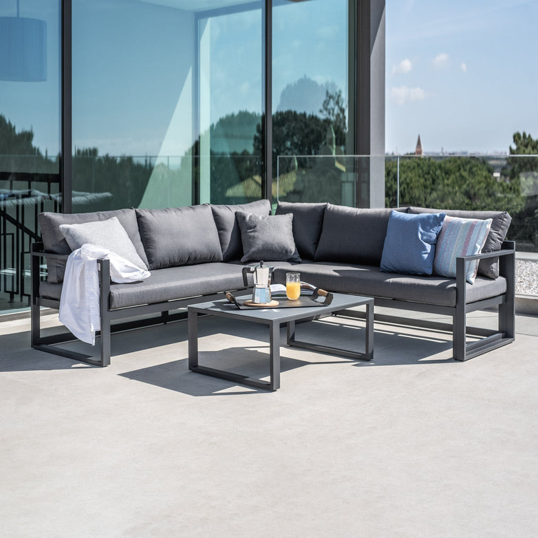 Outdoor living space featuring the Havana Corner Group Set with Reclining Feature and Coffee Table in Charcoal, offering a cozy sectional sofa amidst a picturesque view of trees and buildings in the background. The reclining feature provides an extra touch of comfort to your ideal patio arrangement.