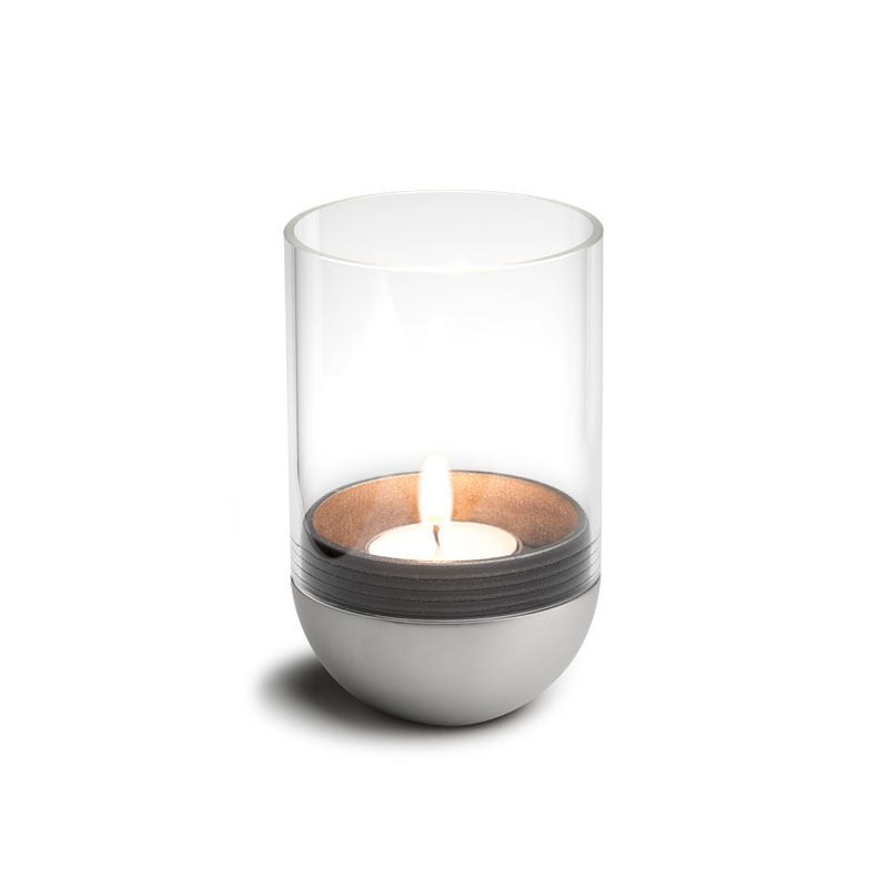 A lit tealight candle in a clear glass holder with a metallic base, reminiscent of the Höfats GRAVITY CANDLE Lantern, perfect for both indoor and outdoor ambiance.