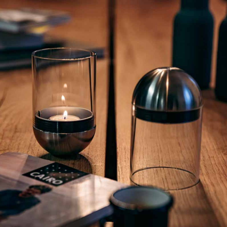 A lit Höfats GRAVITY CANDLE Lantern in a glass holder next to a metal-topped glass cover on a wooden table with a magazine nearby.