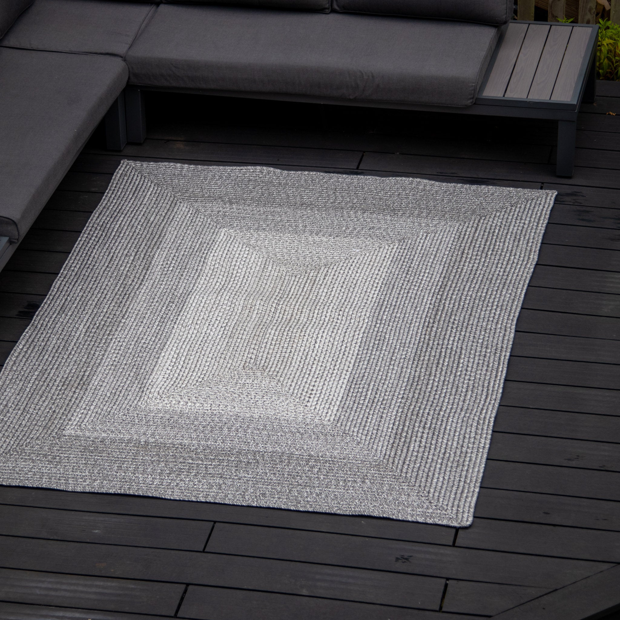 Outdoor space with a Hestia 220cm x 160cm Weatherproof Indoor and Outdoor Rug in Walnut Grey, featuring a white geometric pattern, on a dark wooden deck, surrounded by grey seating.