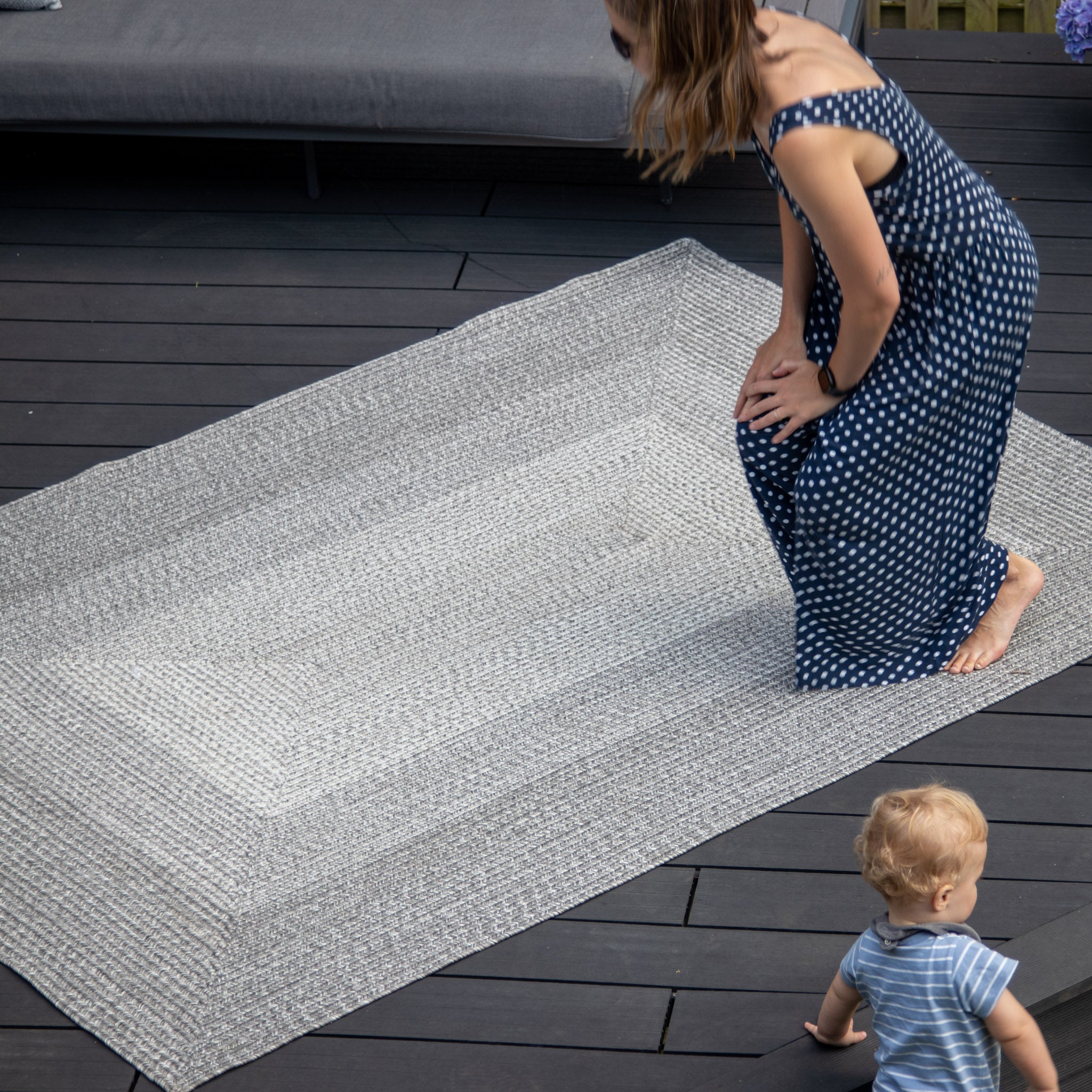A woman in a blue polka dot dress straightens the Hestia 220cm x 160cm Weatherproof Indoor and Outdoor Rug in Walnut Grey on a wooden deck while a toddler in a striped outfit looks on, adding a touch of charm to the peaceful scene with its UV-resistant features.