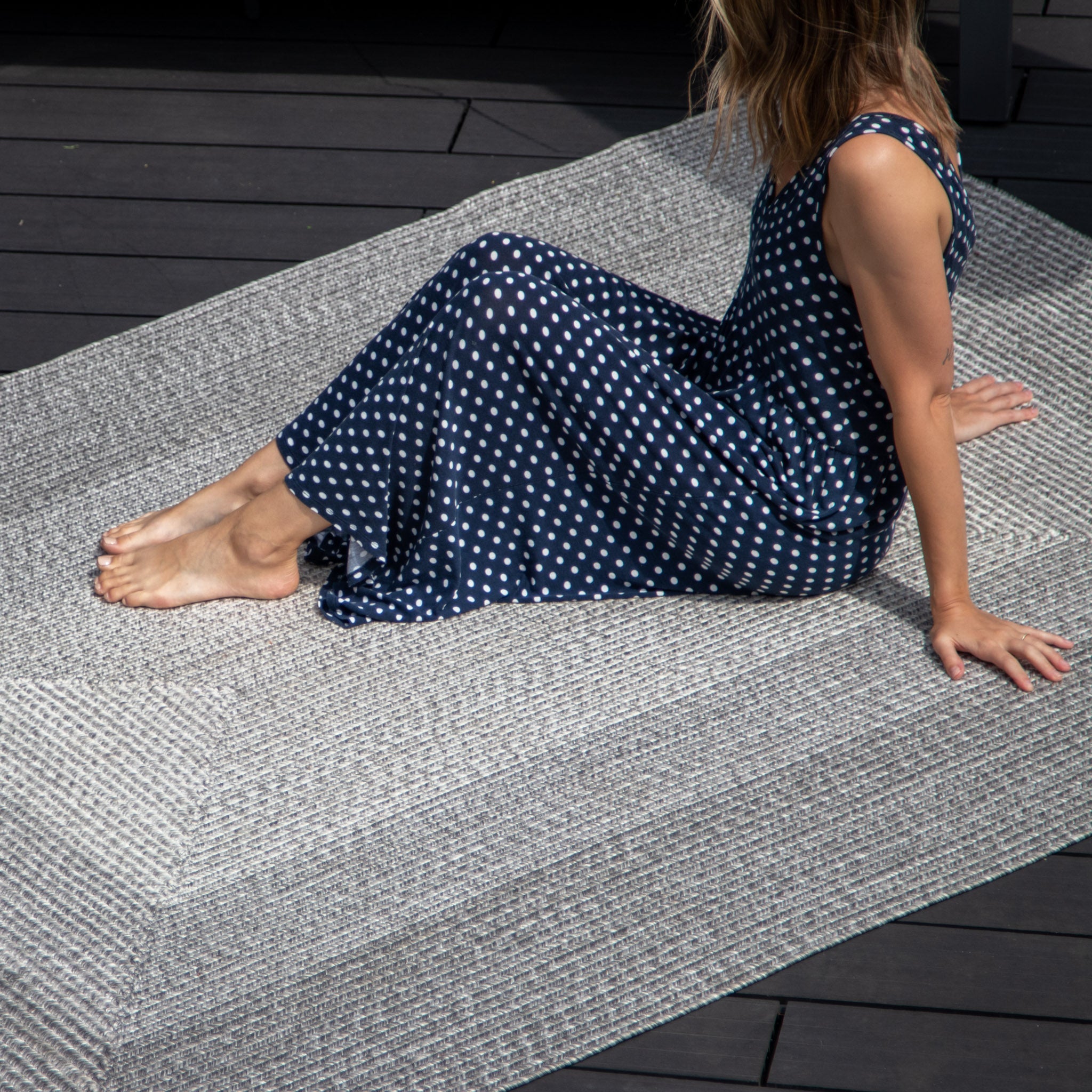 Person in a navy polka dot dress sits on a Hestia 220cm x 160cm Weatherproof Indoor and Outdoor Rug in Walnut Grey on a wooden deck, with their legs relaxed and hair partially covering face.
