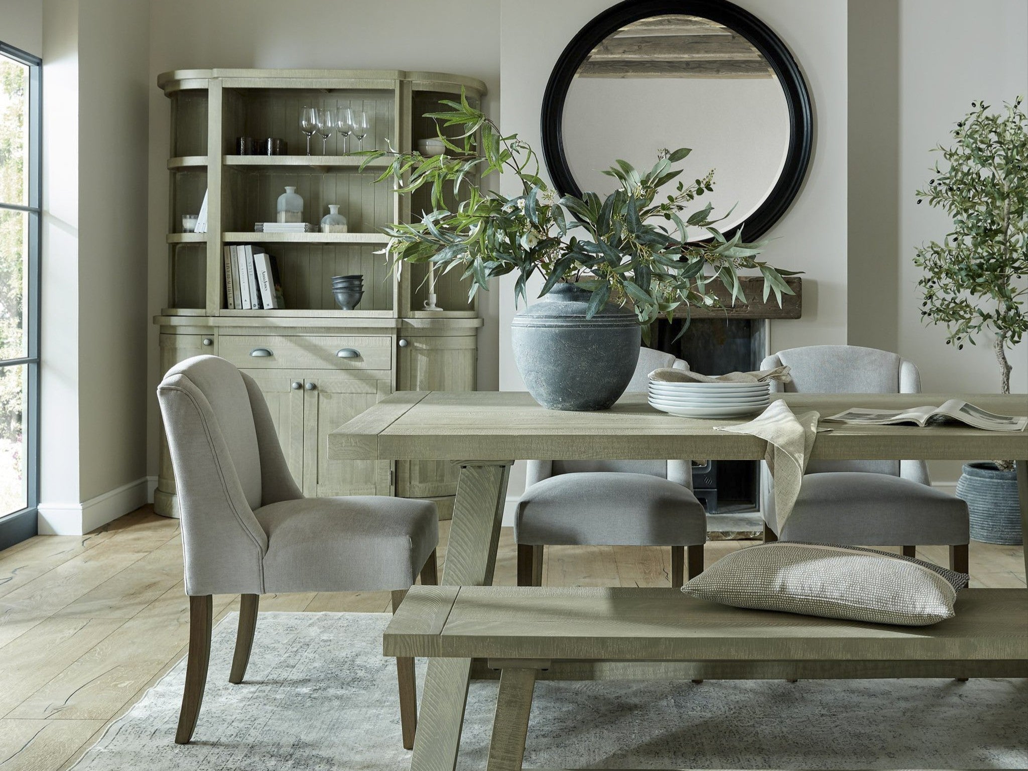 Sage green dining table and bench paired with upholstered dining chairs, set beside a matching glass-fronted display cabinet, styled with a round mirror, stone vase, and greenery for a relaxed country-modern dining room.