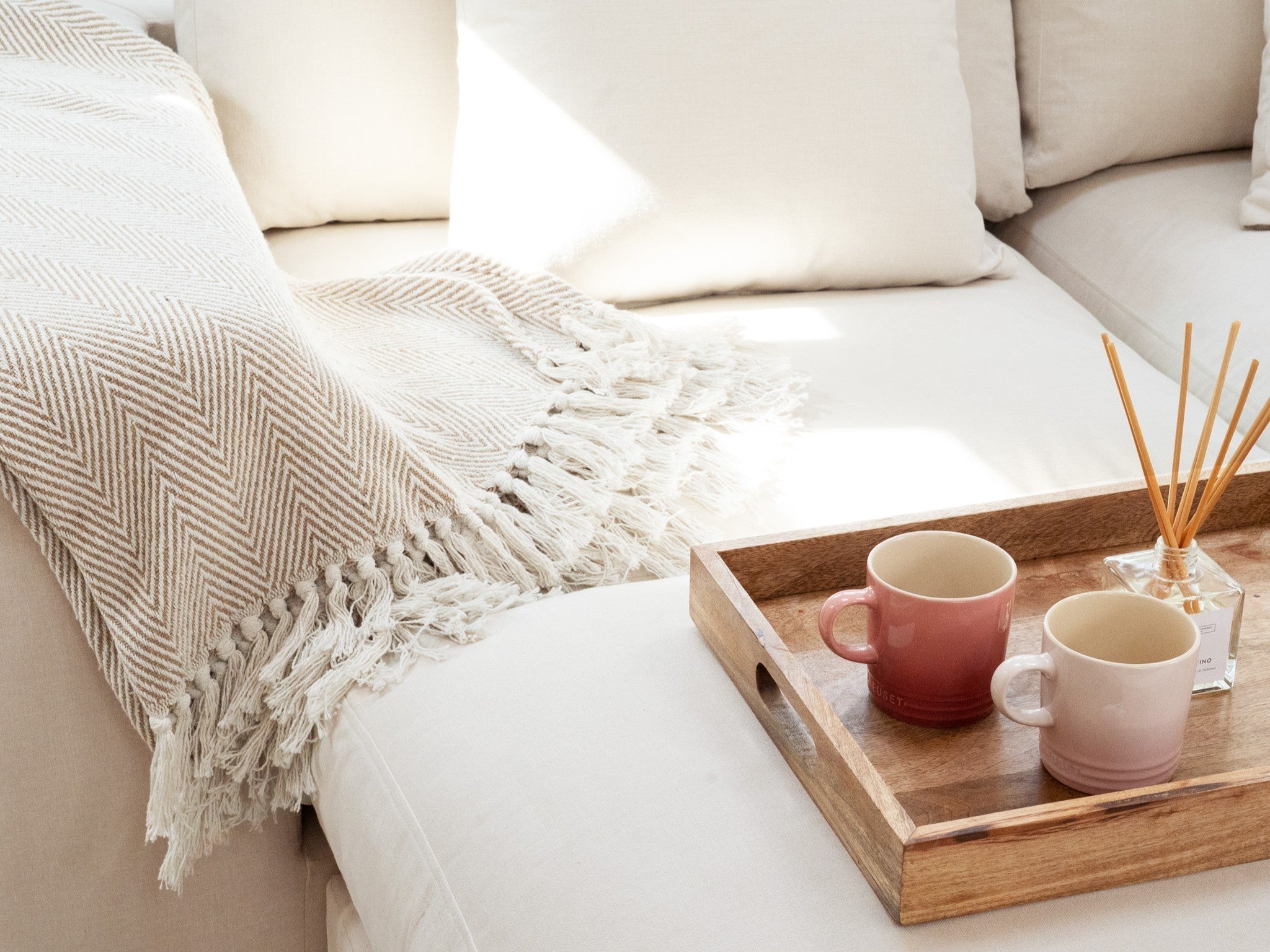 Beige herringbone-weave throw with tasselled fringe draped over a cream sofa arm, styled with a wooden tray holding a reed diffuser and pink Le Creuset mugs.