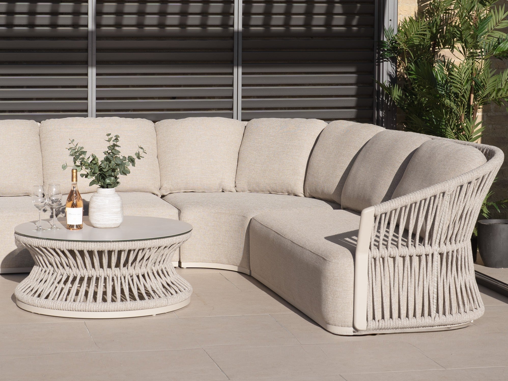 Beige corner sofa set with woven rope sides and an oval rope coffee table, styled with wine glasses and eucalyptus on a sunlit patio.