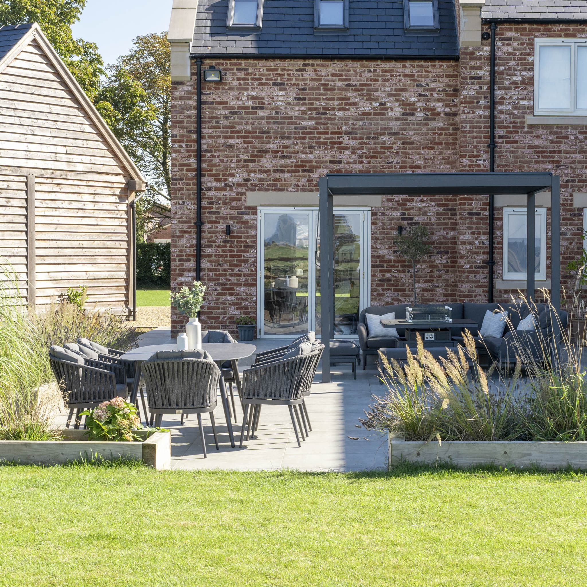 A pergola with furniture underneath it and an 8 seat dining set next to this on a light grey patio with a house behind it.