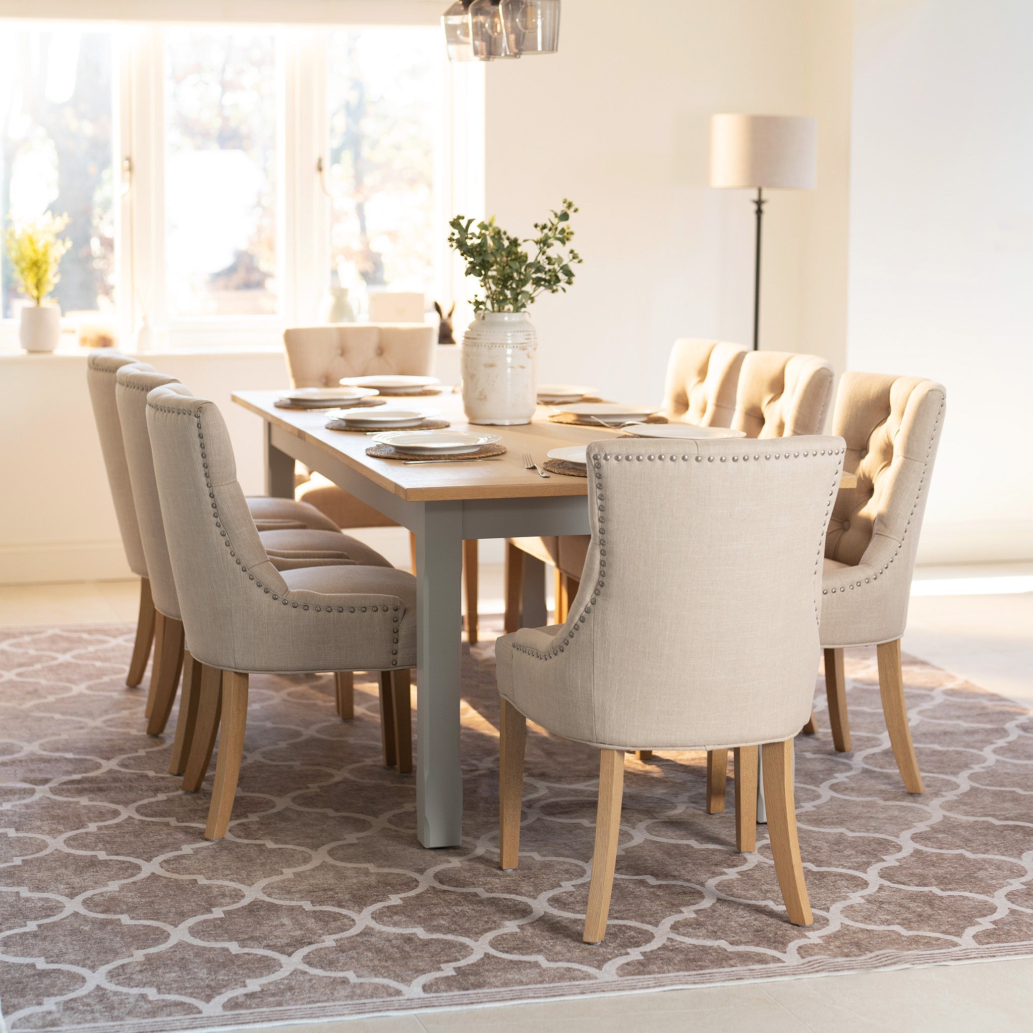 Dining room with a rustic table, beige upholstered chairs, and a potted plant centerpiece on a patterned rug.