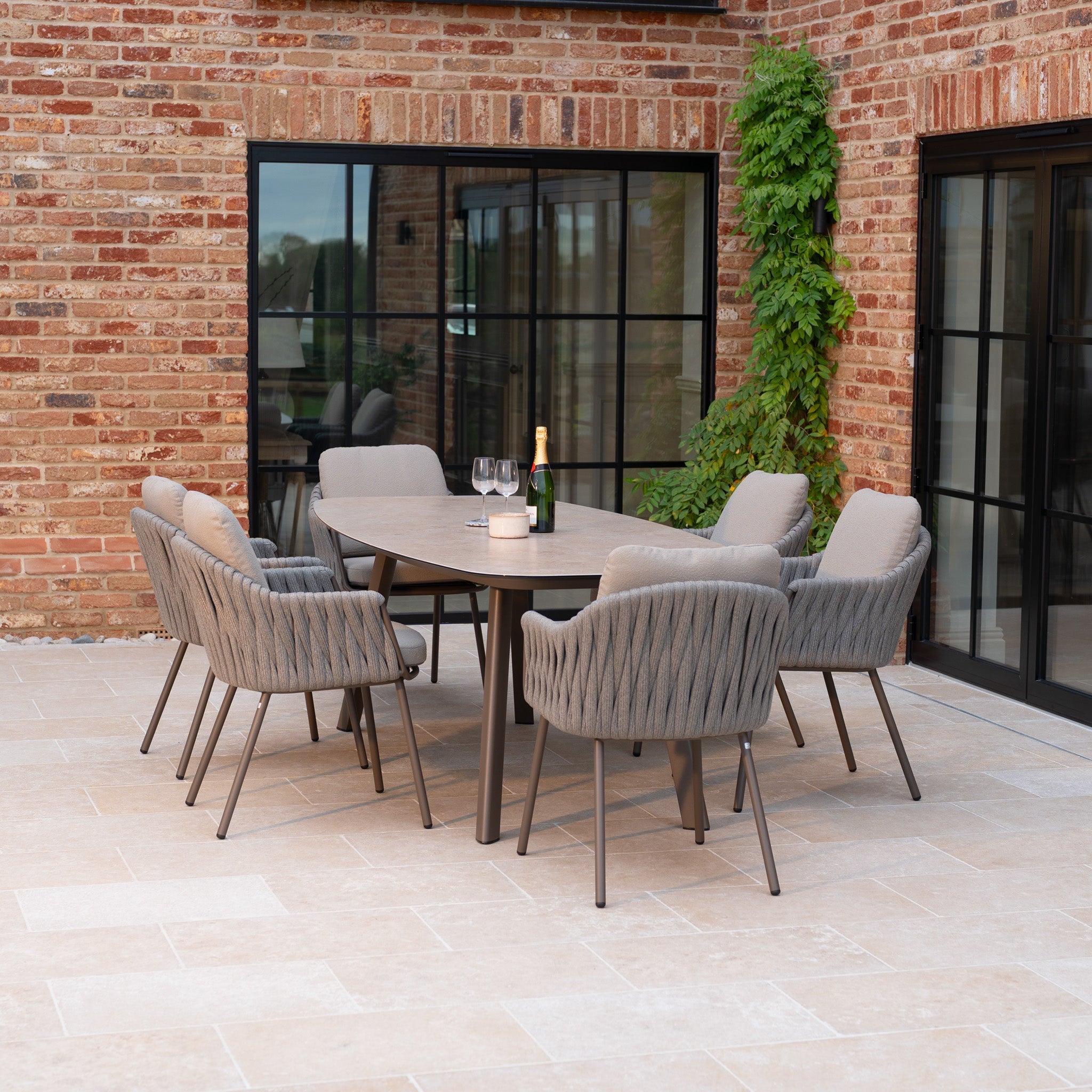 Modern outdoor dining set with six cushioned chairs and a table, set on a patio beside a brick building.