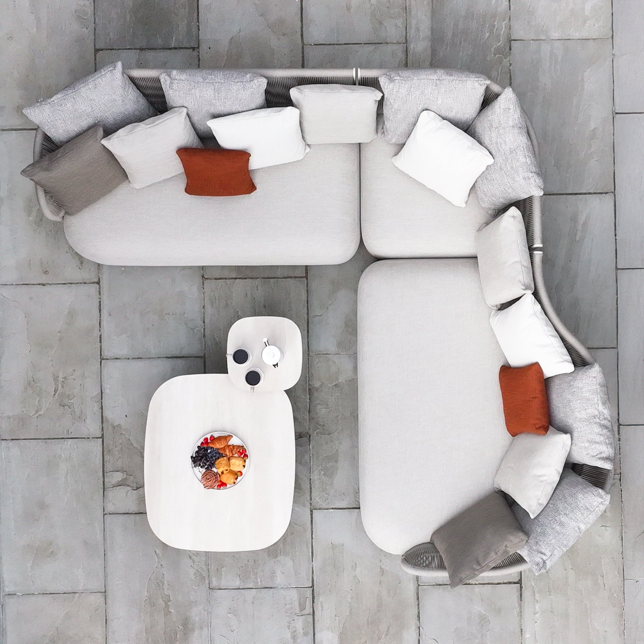 Overhead view of an L-shaped sofa with assorted pillows, two tables, and a fruit platter on a stone floor.