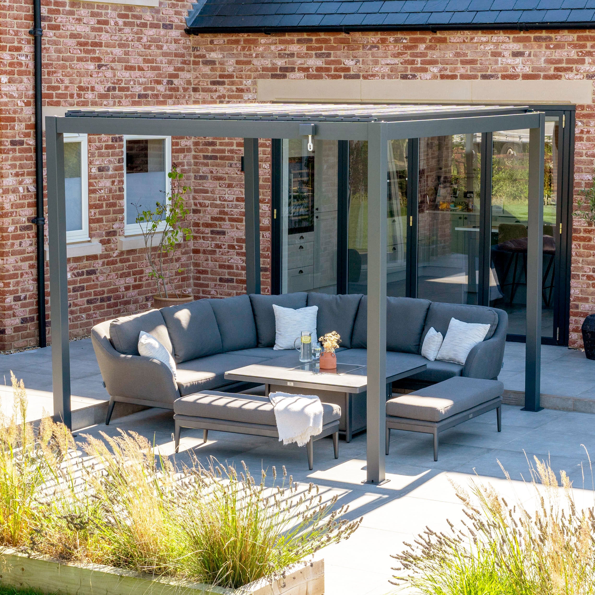 Modern outdoor seating area with grey sofa set under a pergola, next to a brick house with large glass doors.