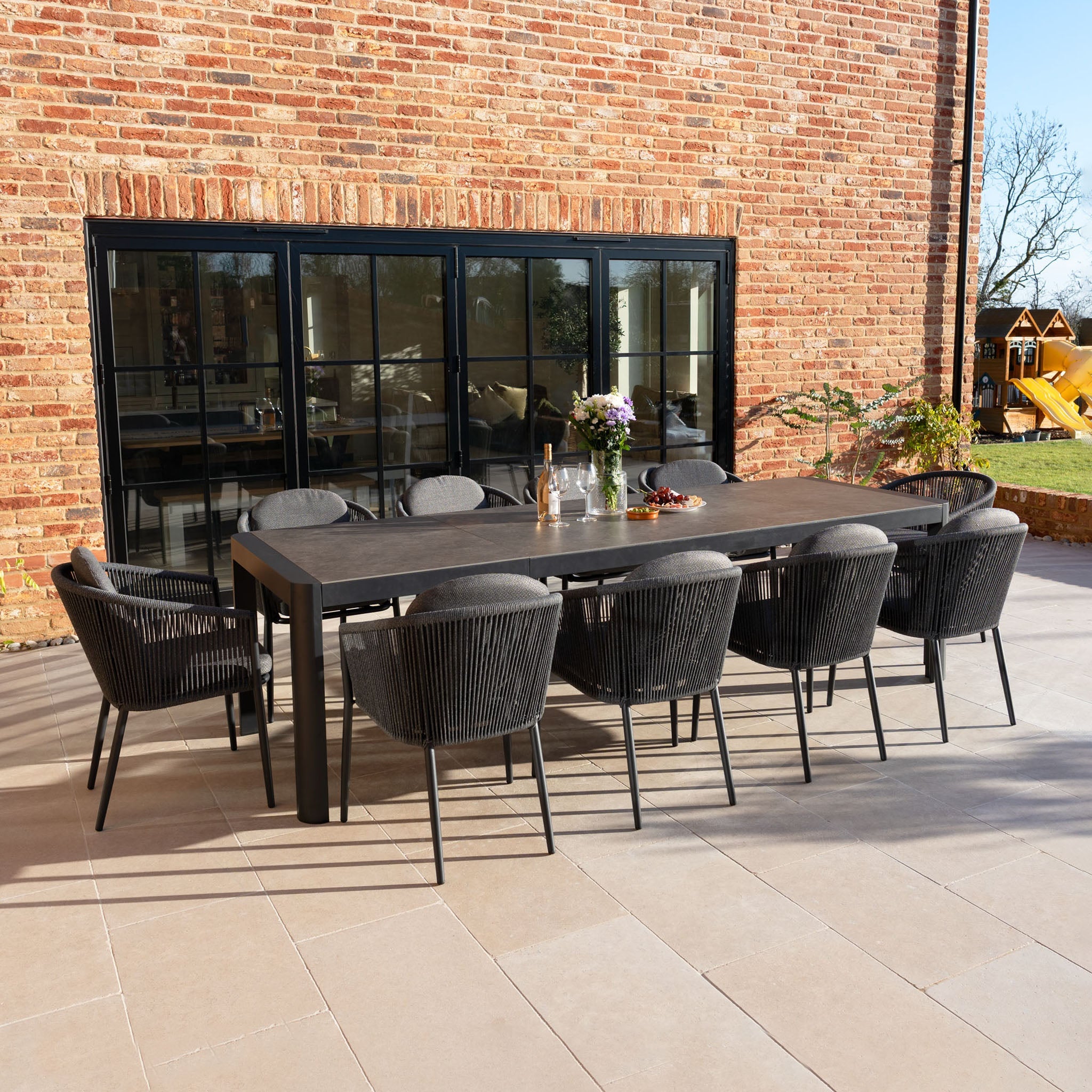 Modern outdoor dining table with eight black chairs on a patio next to a brick wall with large windows.