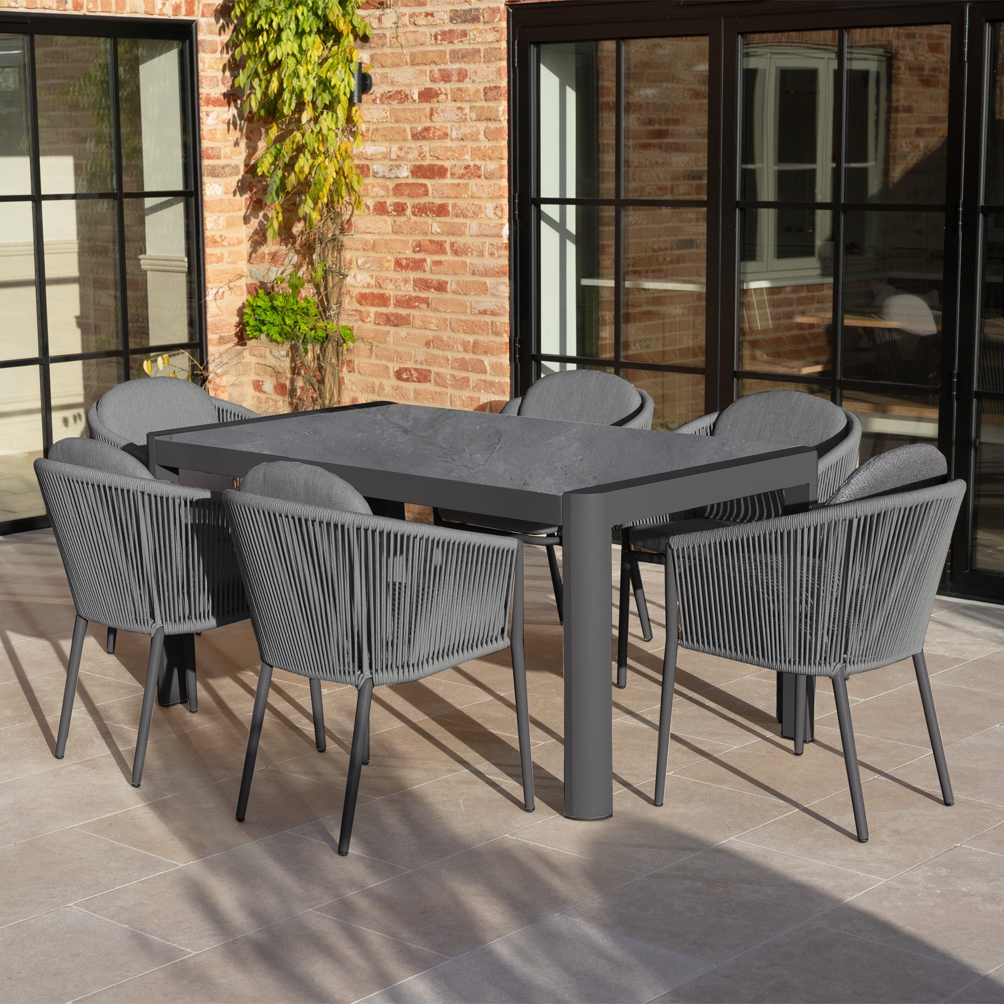 Modern gray outdoor dining set with eight chairs on a tiled patio beside brick walls and glass doors.