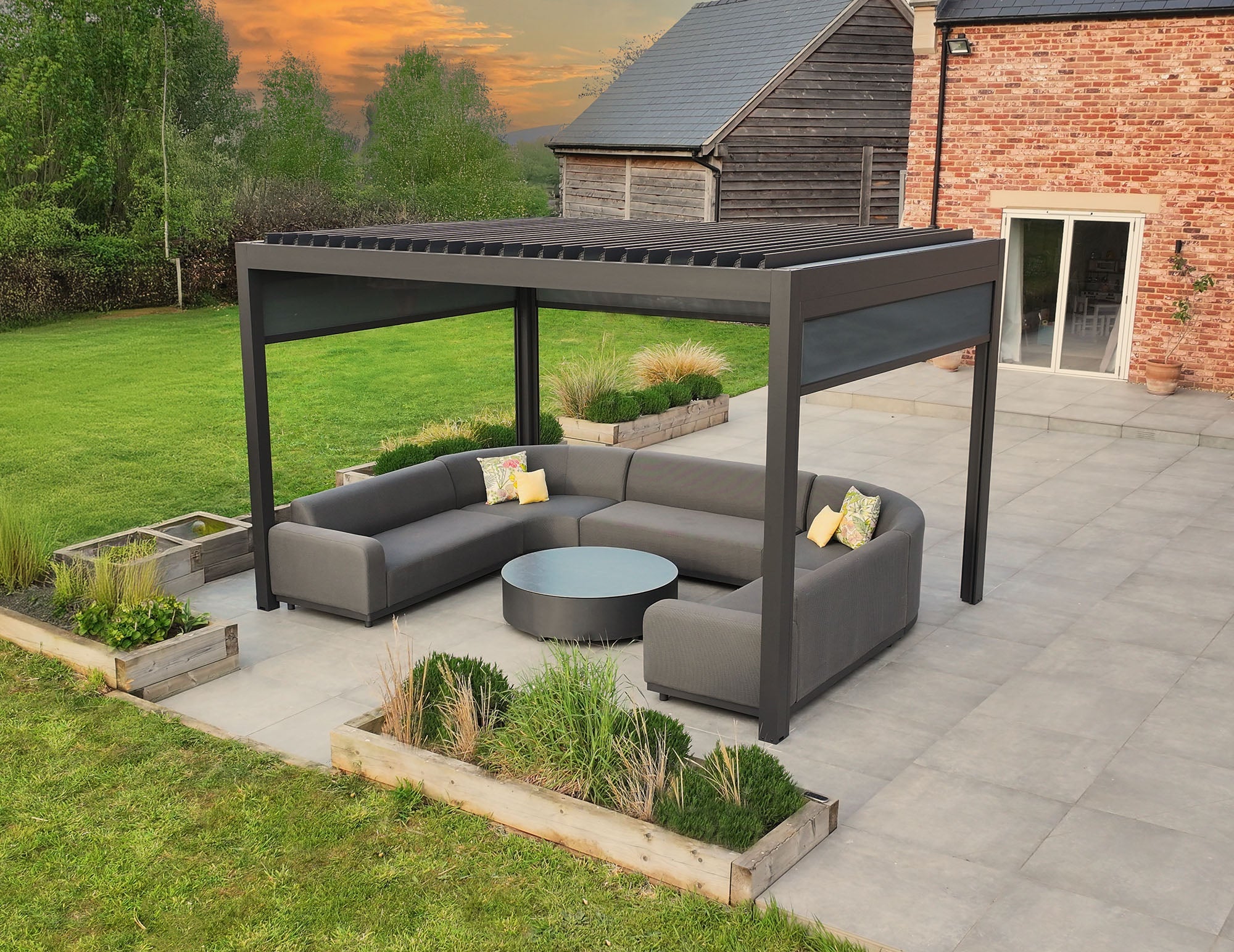 Modern outdoor patio with a large pergola, grey sofas, round table, and raised garden beds at sunset.