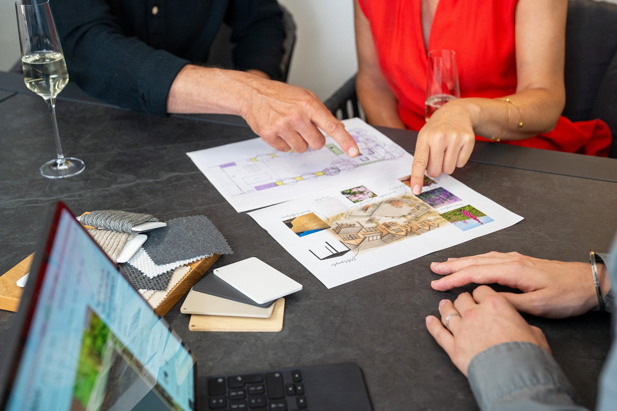 Three people discussing architectural plans at a table with fabric samples, laptop, and drinks.
