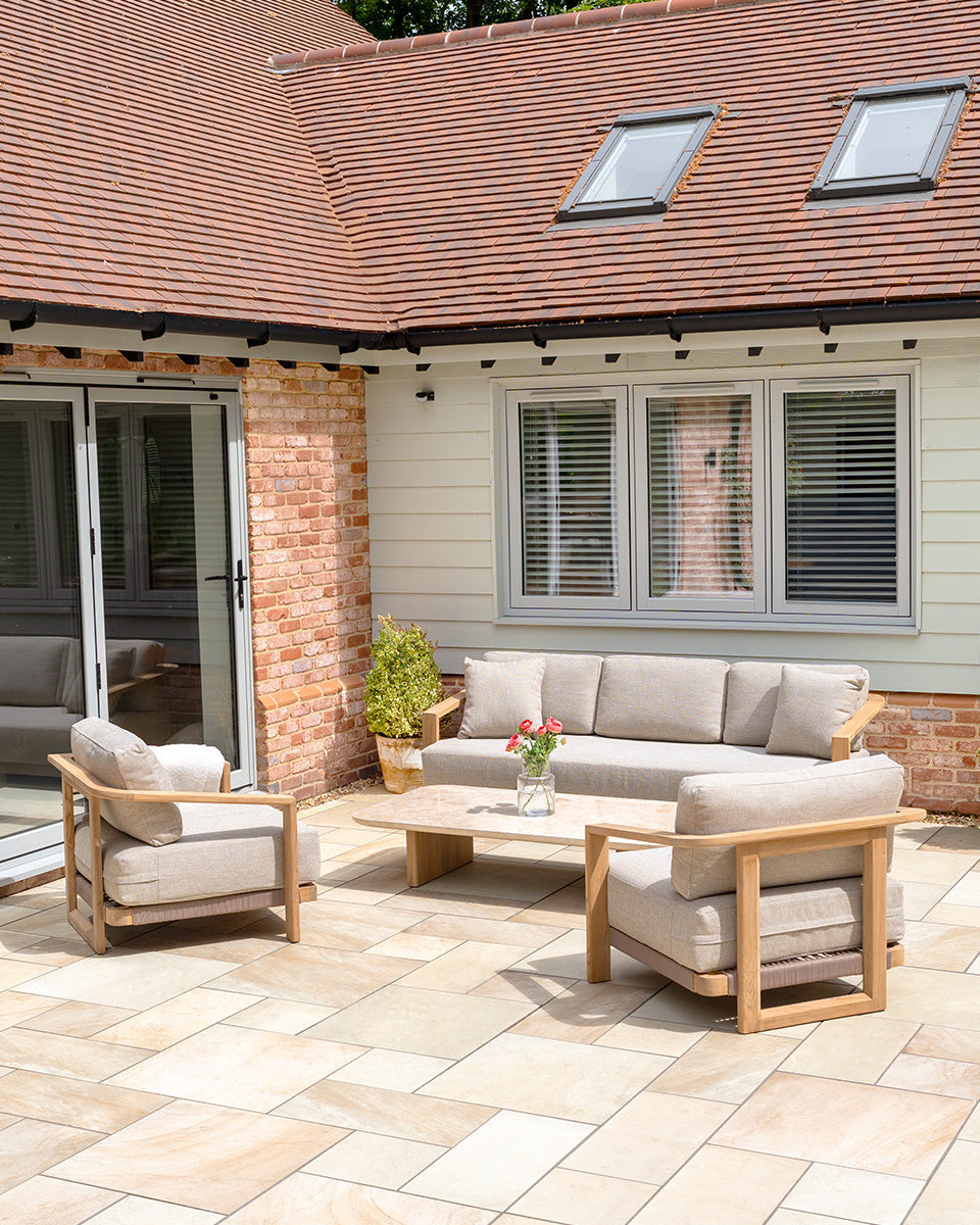 Patio with beige outdoor furniture, a coffee table with flowers, and a brick house with large windows.
