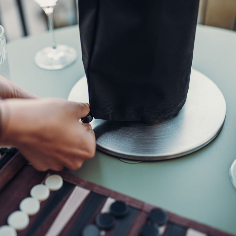 Hands adjusting the Höfats GRAVITY CANDLE Base, featuring a black cover on a stainless steel base, with a backgammon game and drinks in the background.