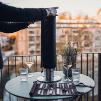 A person setting up a device on a balcony table with backgammon, wine glasses, and a scenic background ensures storage protection with the Höfats SPIN 90 & 900 Cover.