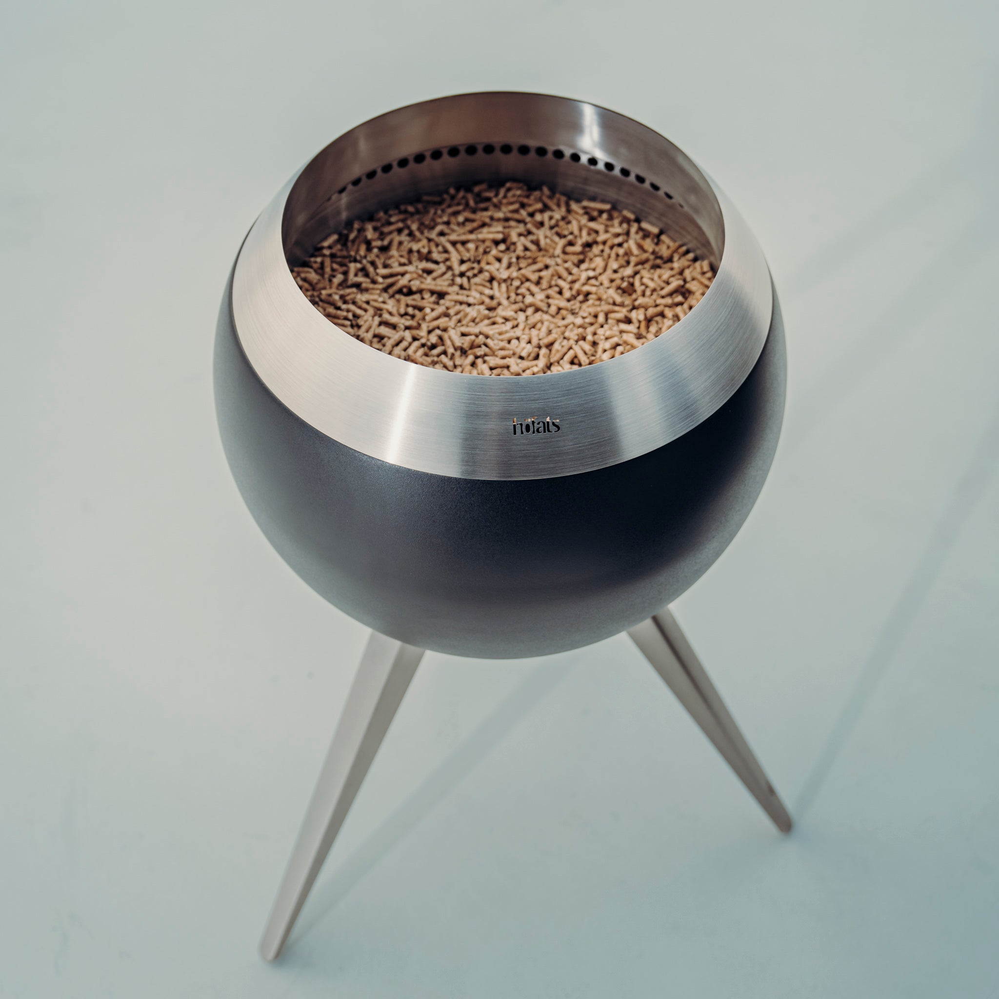 A contemporary Höfats MOON 45 Fire Bowl with a low stand, designed with a spherical shape and supported by three metallic legs, filled with wood pellets and evoking the essence of black MOON fire art, displayed against a plain background.