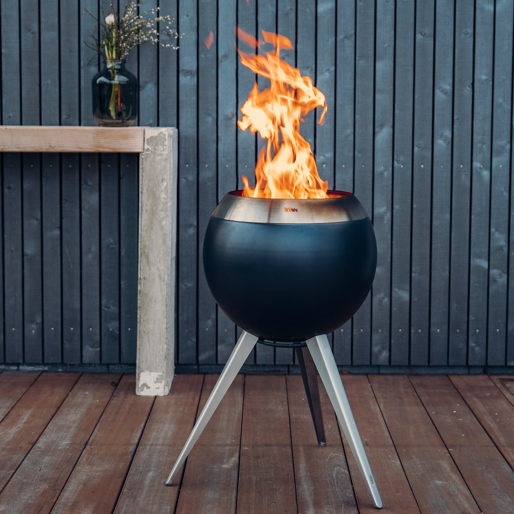 A round, modern Höfats MOON 45 Fire Bowl with High Stand adorned with flames burning, set on a wooden deck near a table with a vase of flowers, showcasing revolutionary combustion technology for an efficient burn.