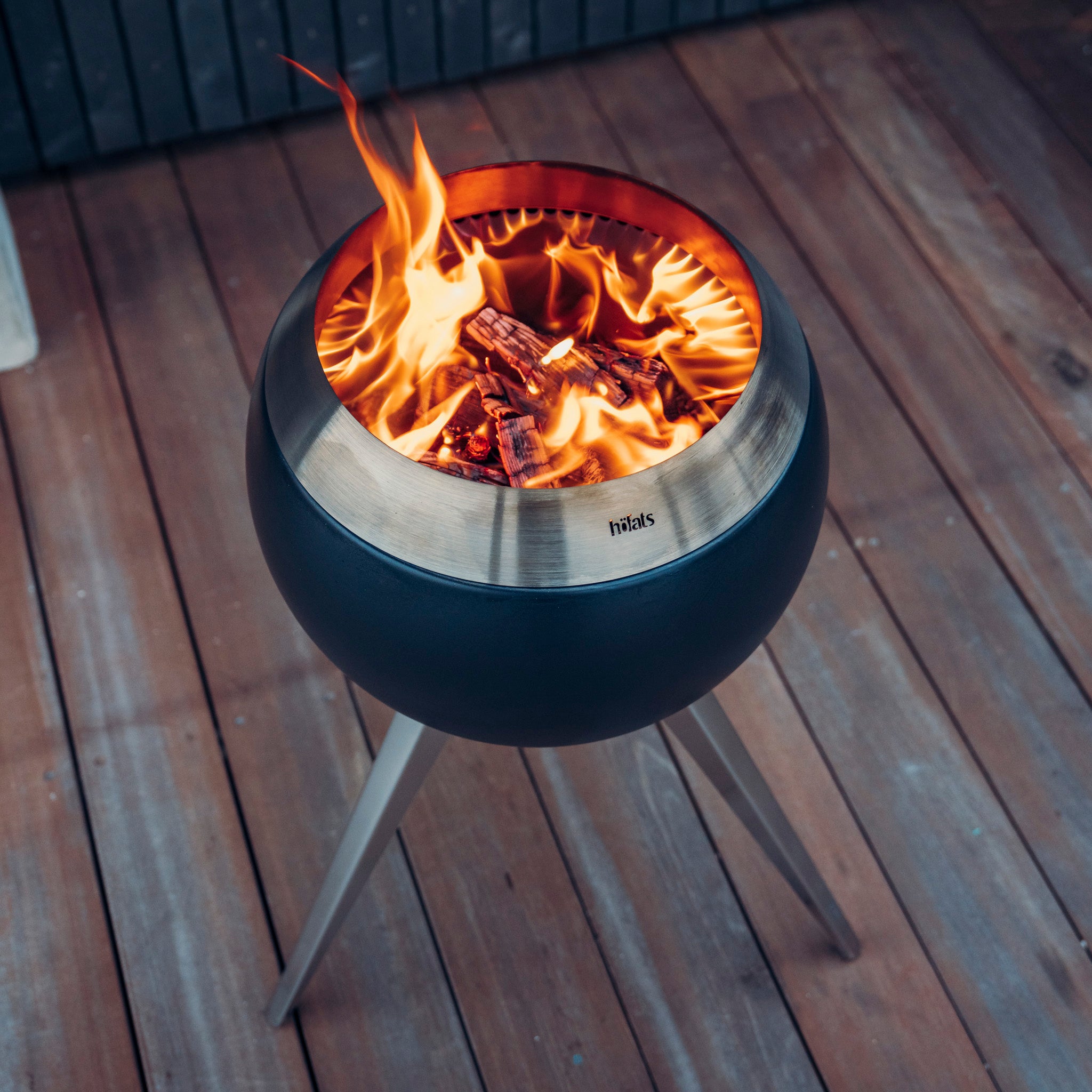 A modern, metal fire pit showcasing a spherical design on a wooden deck features revolutionary combustion technology that enhances the burning wood and flames—a perfect description of the Höfats MOON 45 Fire Bowl with High Stand.