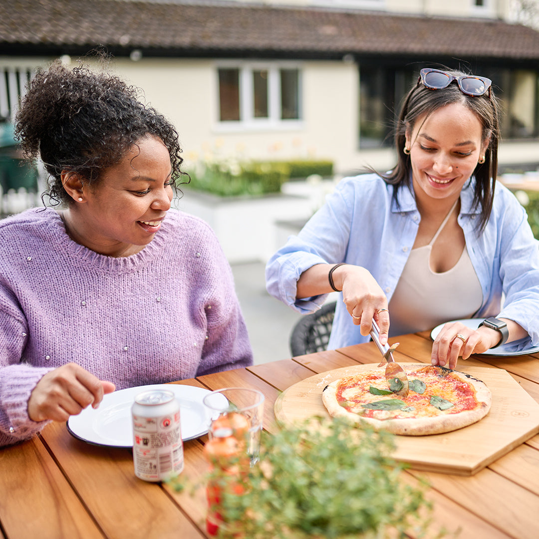 Two women smile and sit at a wooden table outside, one joyfully cutting a pizza on a sleek 16" Bamboo Pizza Board.