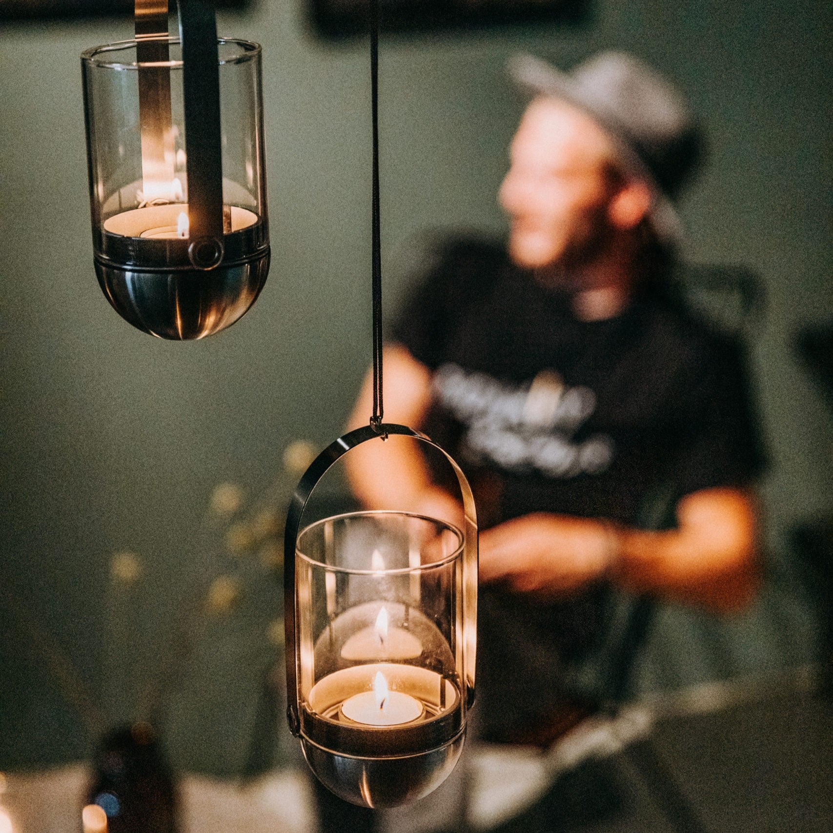 Two Höfats GRAVITY CANDLE Hangers with lit candles; a blurred person wearing a hat in the background, all exquisitely crafted from brushed solid stainless steel.
