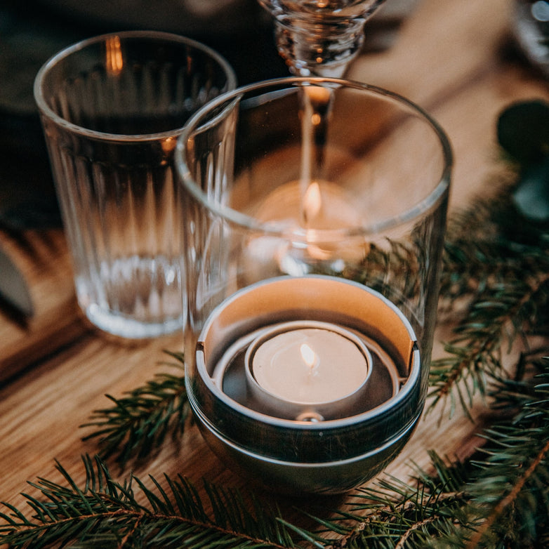 A Höfats GRAVITY CANDLE Lantern sits elegantly in a glass holder on a wooden table with greenery, beside an empty water glass and a wine glass.