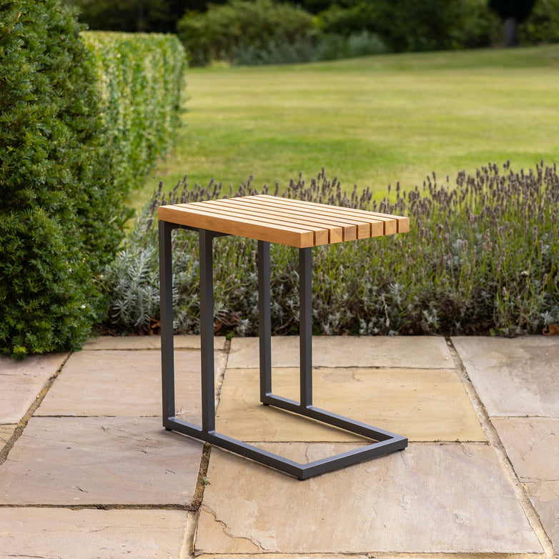 The Cube Side Table with Teak Top in Charcoal showcases its modern design on a stone patio, complemented by a lush garden backdrop.