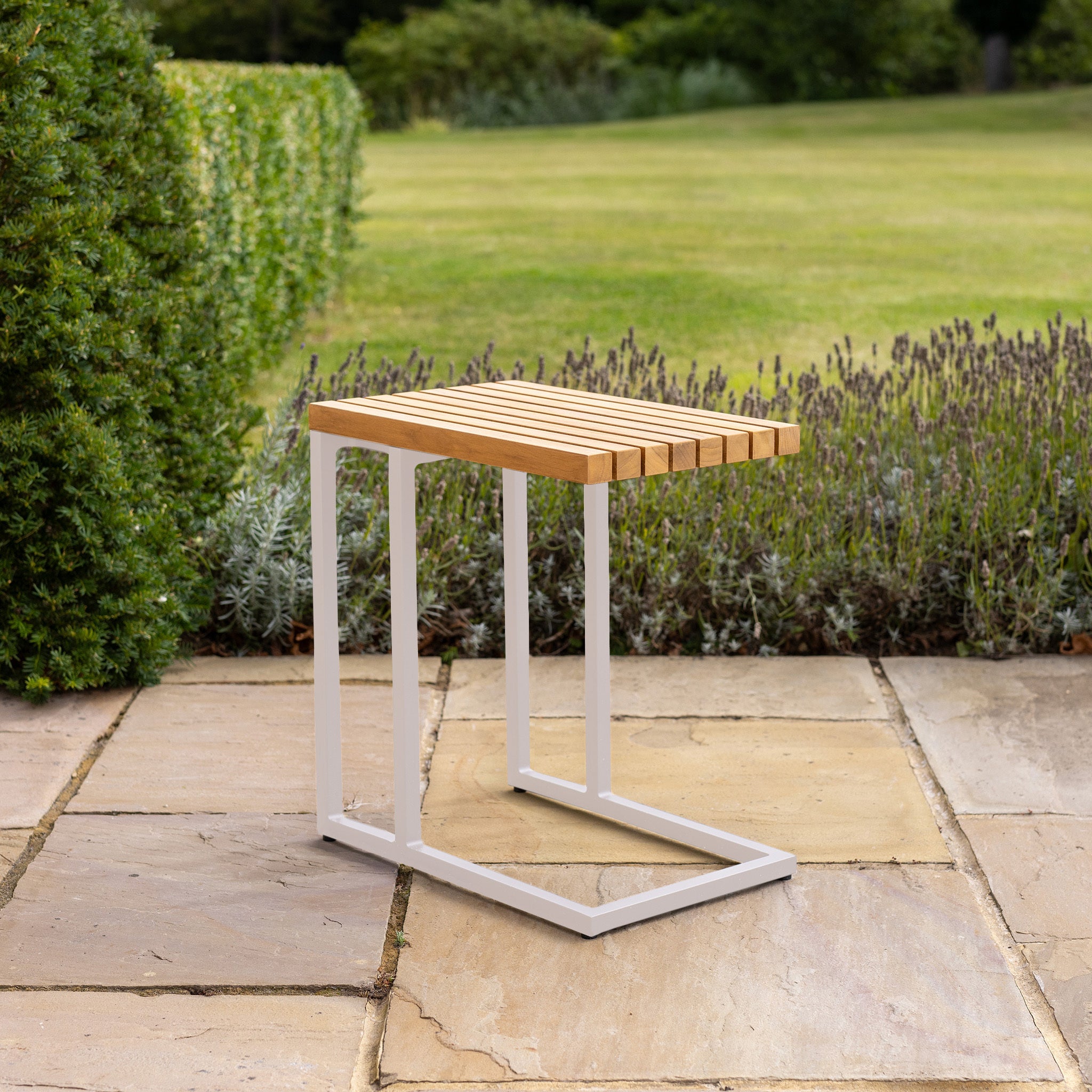 The Cube Side Table with Teak Top in Latte sits elegantly on the stone patio, encircled by vibrant greenery and a manicured lawn. Its modern wood-and-metal design seamlessly integrates with nature, while the teak top offers enduring charm.