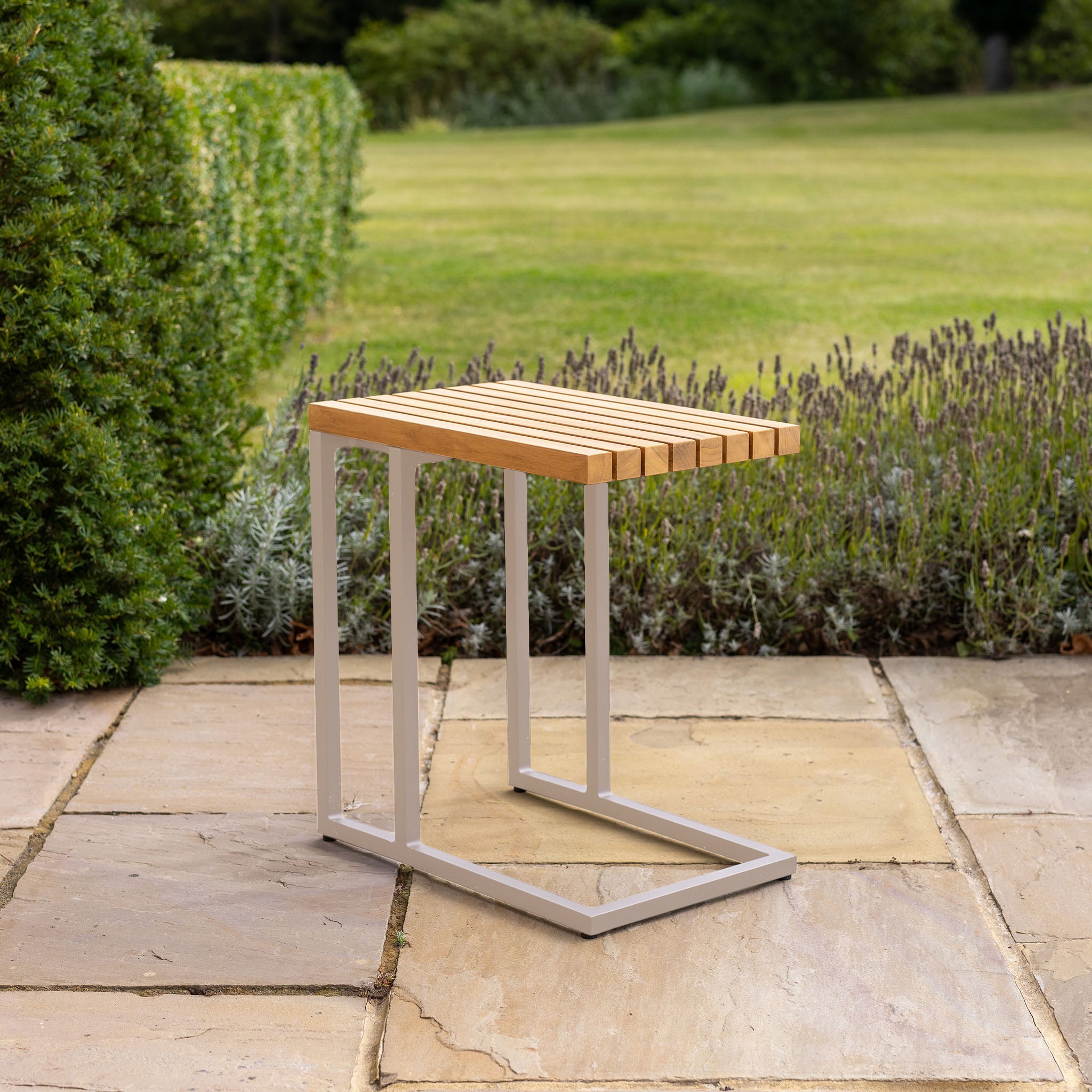 The Cube Side Table with Teak Top in Latte sits on a stone patio, with grass and bushes in the background.