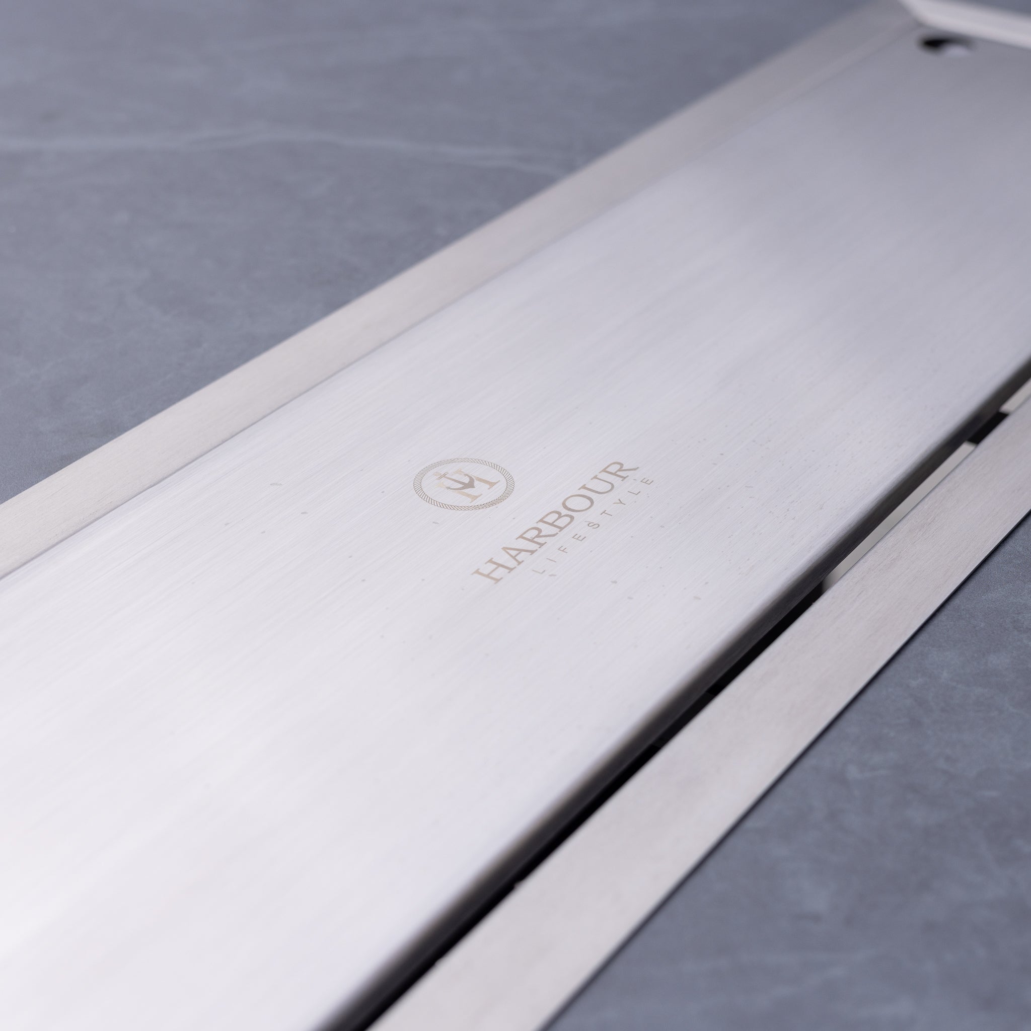 Close-up of a sleek silver product with the "Harbour Lifestyle" logo engraved on it, placed on a smooth grey surface, next to our Luna 8 Seat Outdoor Fabric Rectangular Ceramic Firepit Dining Set in Grey made of all-weather material.