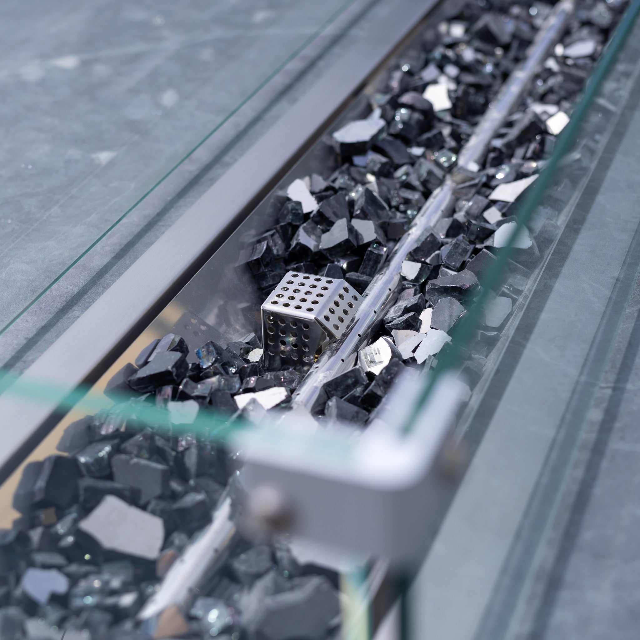 A metal cube nestled among shiny black rocks in a glass display case complements the Luna 8 Seat Outdoor Fabric Rectangular Ceramic Firepit Dining Set in Grey.